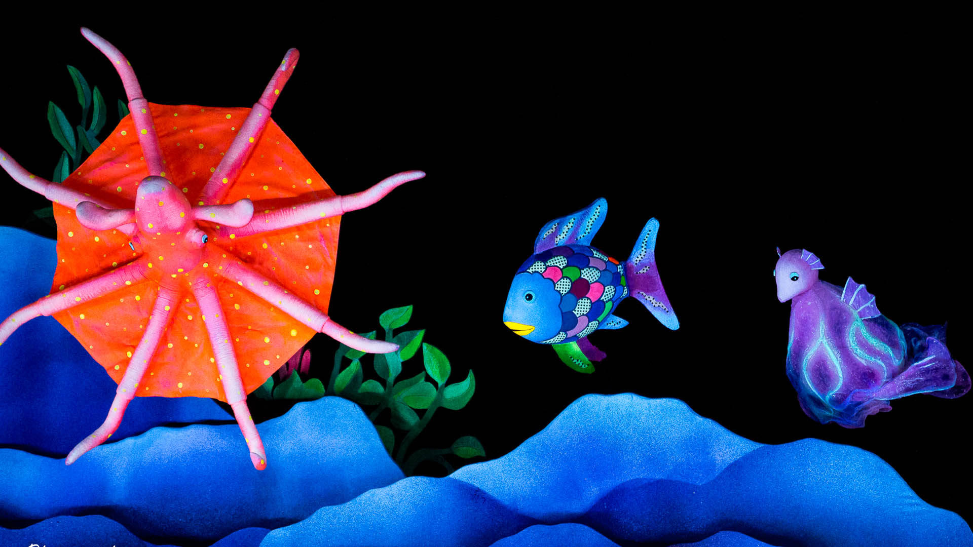 Puppets of a red octopus, purple seahorse and a fish with rainbow scales swimming. The are above blue bedrock and green seaweed.