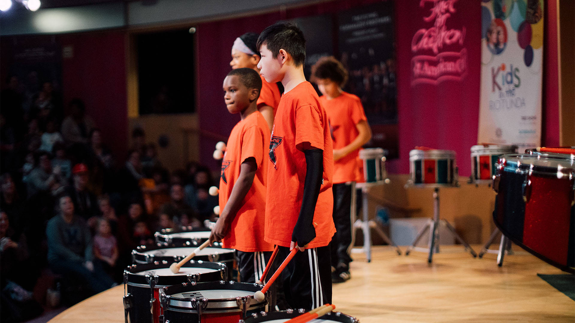 Children in orange shirts standing ready to play marching bass drums on a wooden stage with a crowd in the background