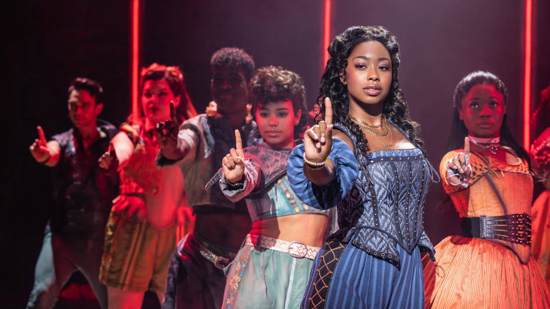 A woman with dark complexion and dark hair in a blue dress and corset holds up her index finger. An ensemble behind her does the same.