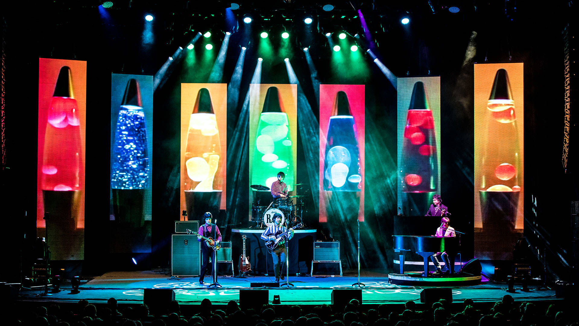A Beatles tribute band performing on stage with guitars, a piano and a drum set. Large lava lamp projections are behind them.