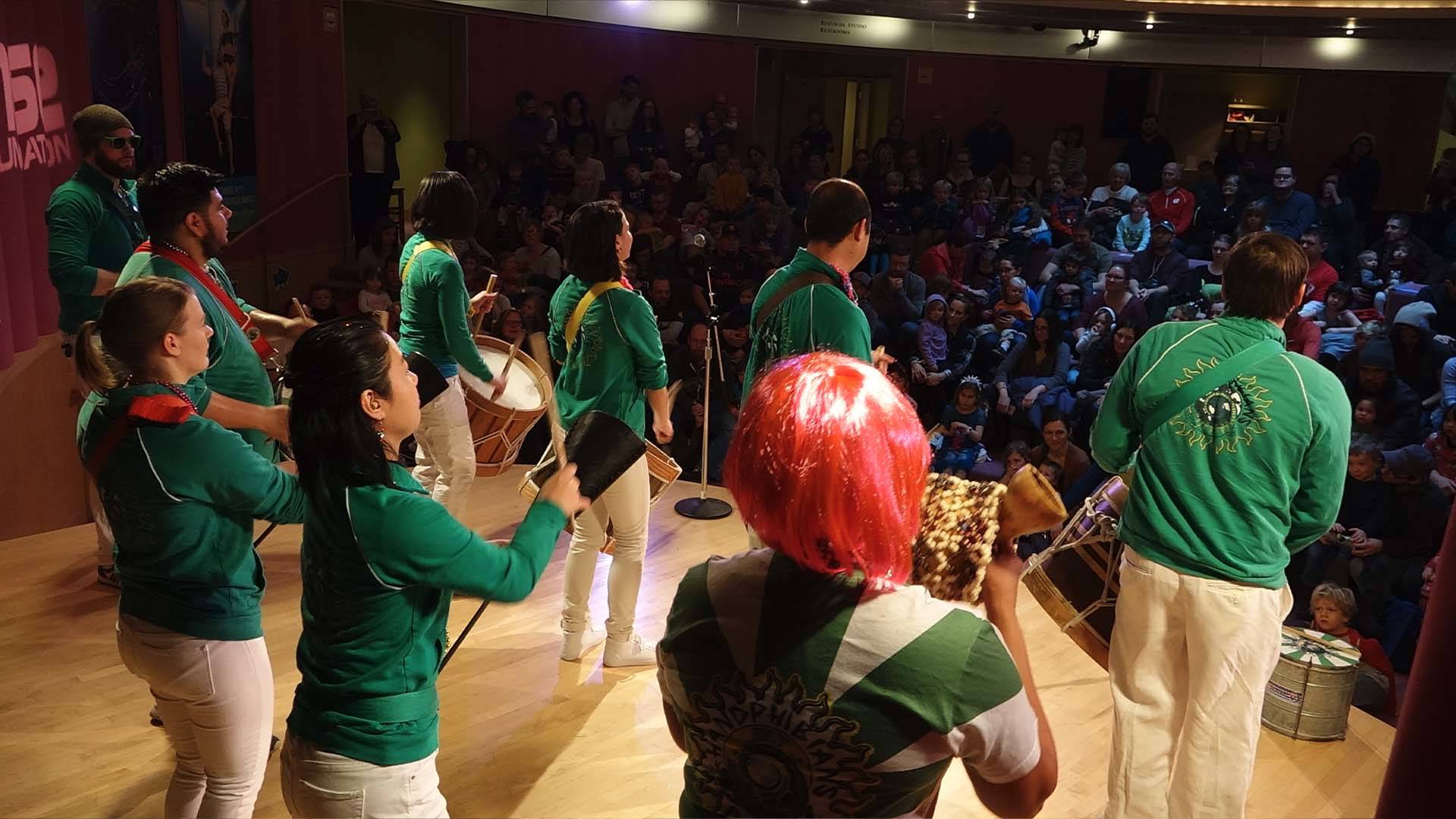A group of musicians in green sweaters play samba instruments. The camera is behind the musicians, looking out towards the audience.