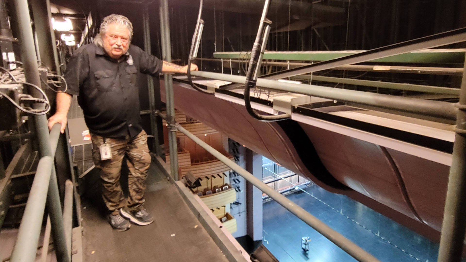 A middle-aged caucasian man with medium length grey hair and a moustache in a black button-down shirt and camoflage pants posing for a photo in one of the catwalks above Overture Hall. The stage is visible far below.