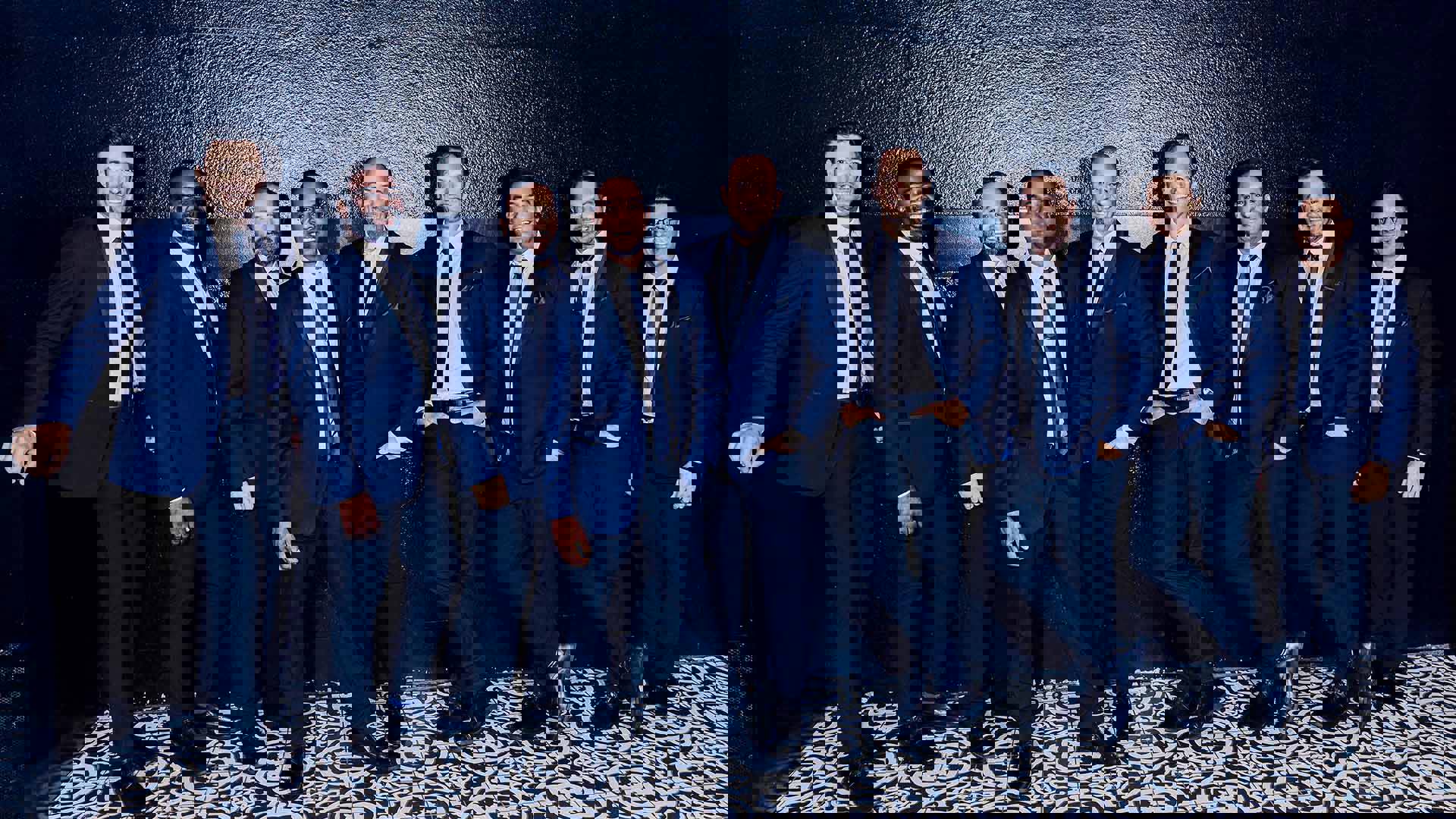 Nine men wearing blue suits standing in front of a black wall.