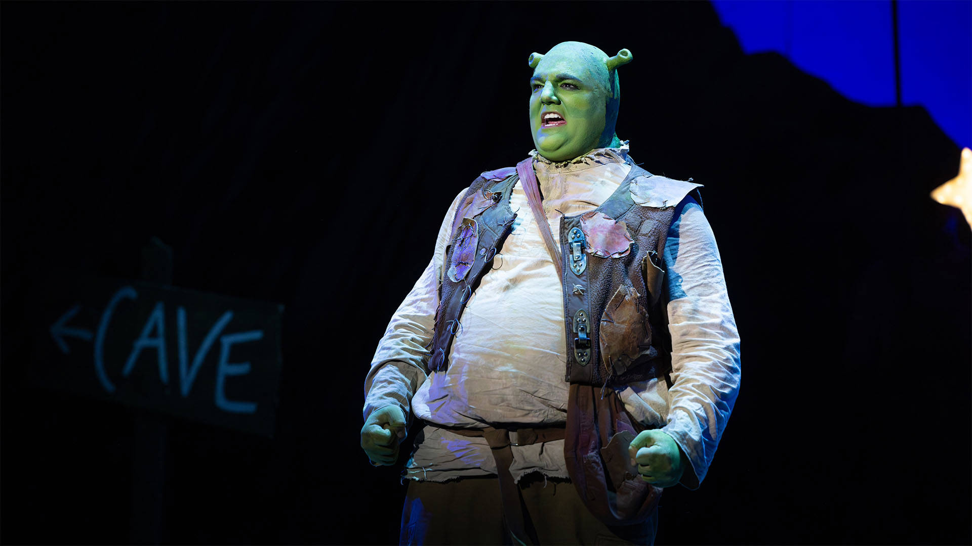 A man with green skin and ogre ears in a hemp shirt, brown belt and patchwork brown vest singing on a dark stage.