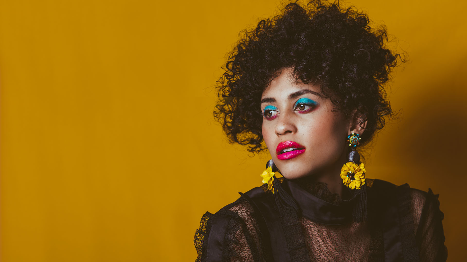 Alea against a golden background. She is wearing her hair up, it is black and very curly. She has distinct makeup - bright blue eye makeup and red lipstick. She is wearing a lacy top and large flowery earings