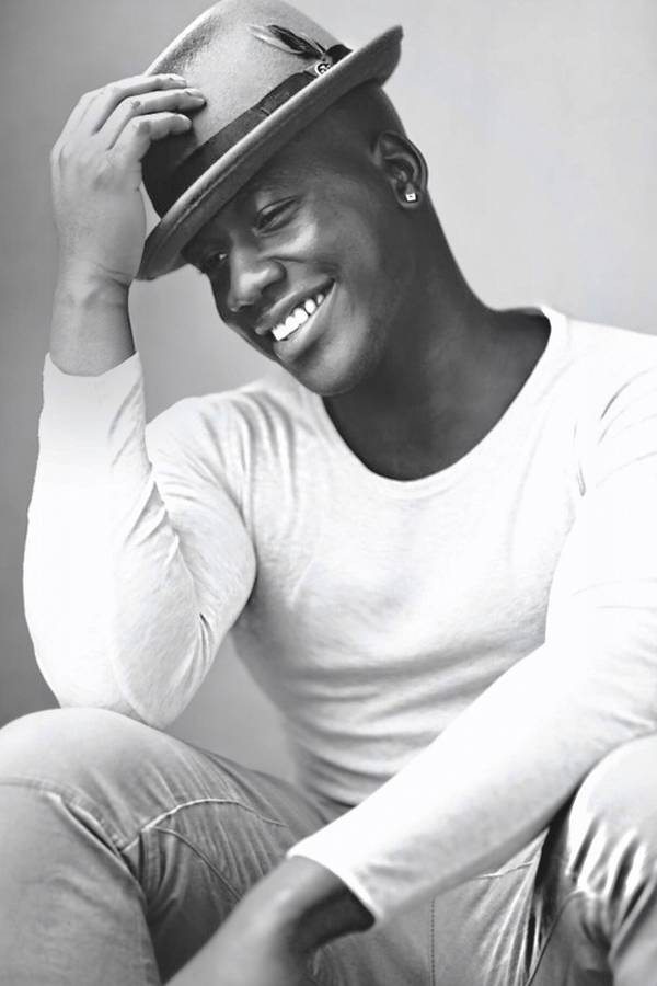 Grayscale photo of a smiling man with dark complexion, dark eyes, a light longsleeve shirt on, earings and a rimmed hat.