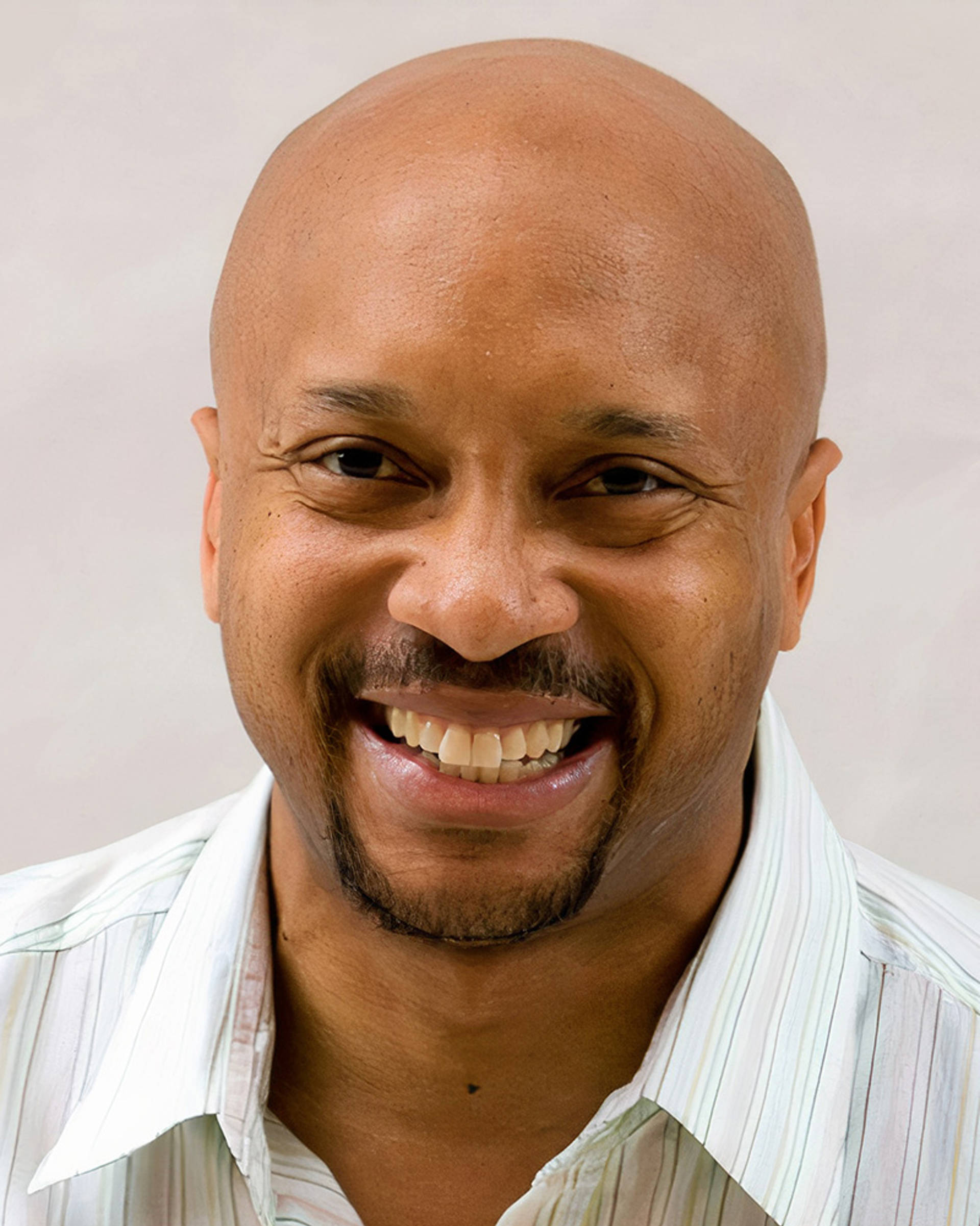 Headshot of an african american man with a bald head, light goatee and dark eyes. He is smiling and wearing a collared shirt.