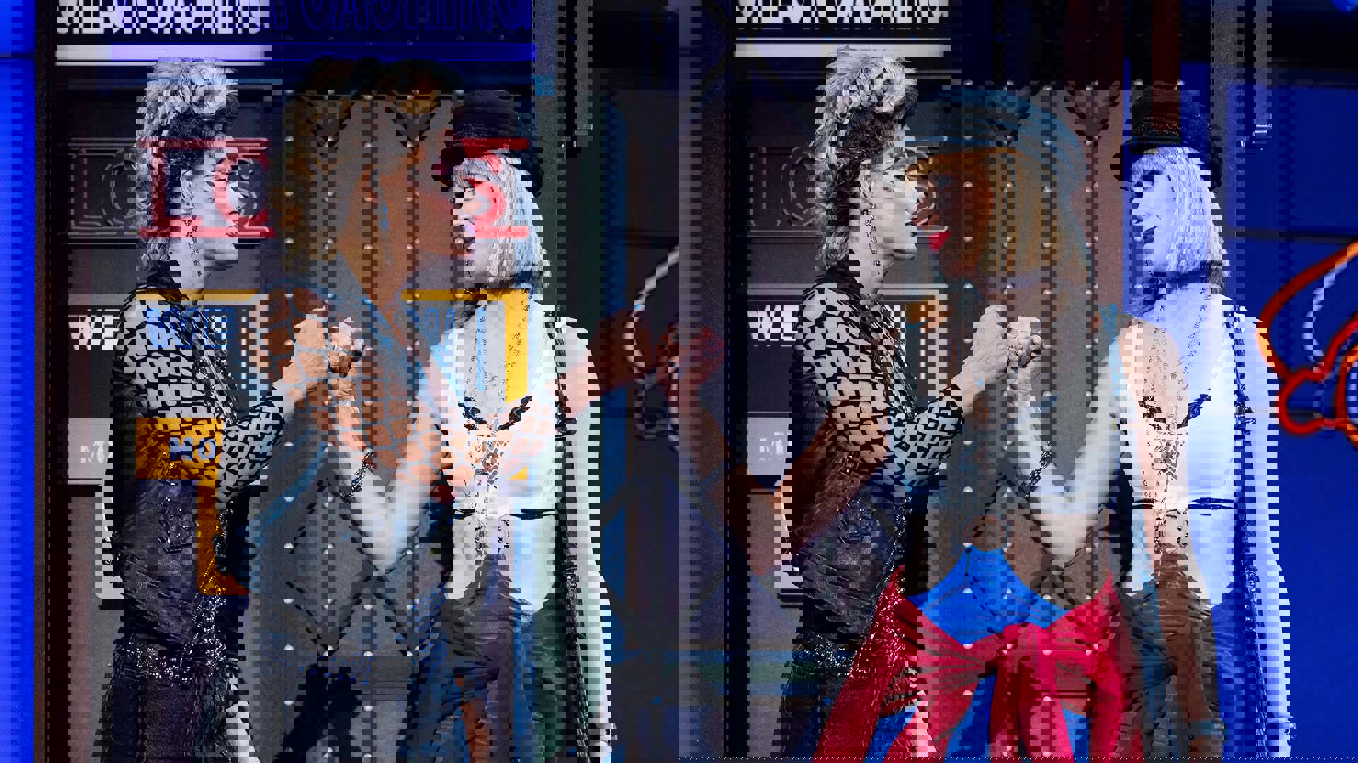 Two women in revealing edgy and revealing outfits hook their pinky fingers and gaze at eachother.