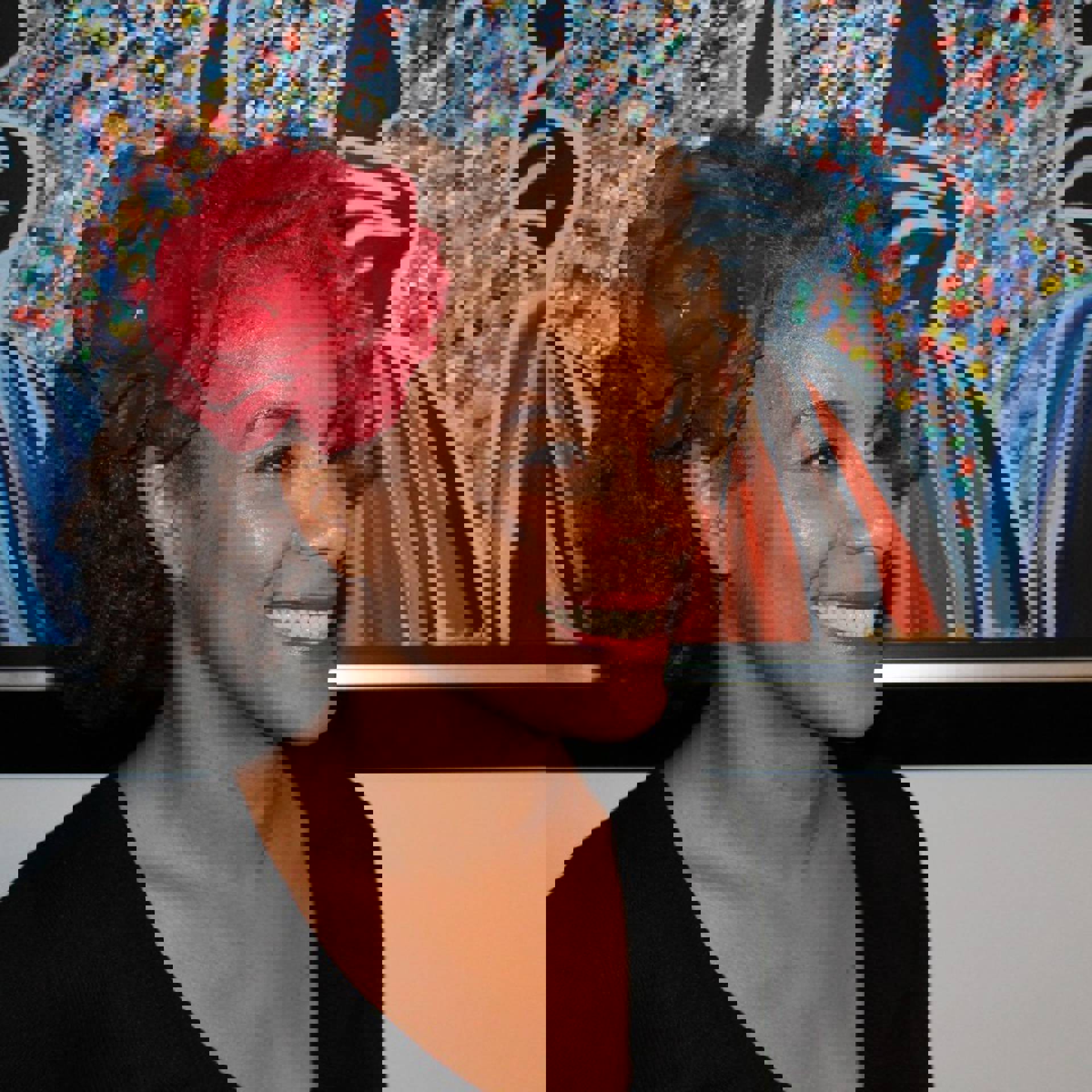 A headshot of a smiling middleaged African American woman with medium length hair, a flower decoration in her hair, and a black top.