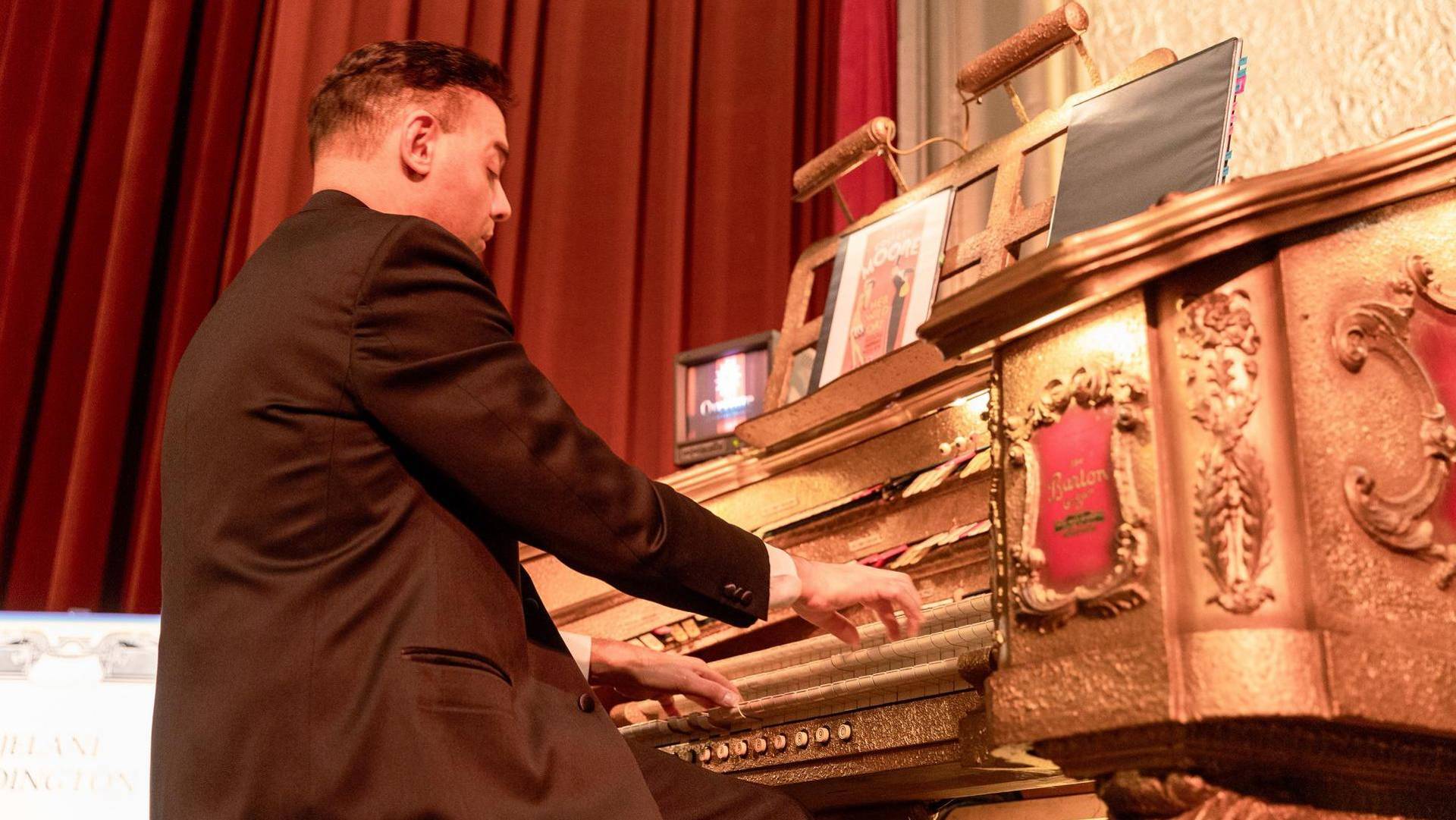 A man in a suit jacket with dark hair and light complexion plays a golden organ console.
