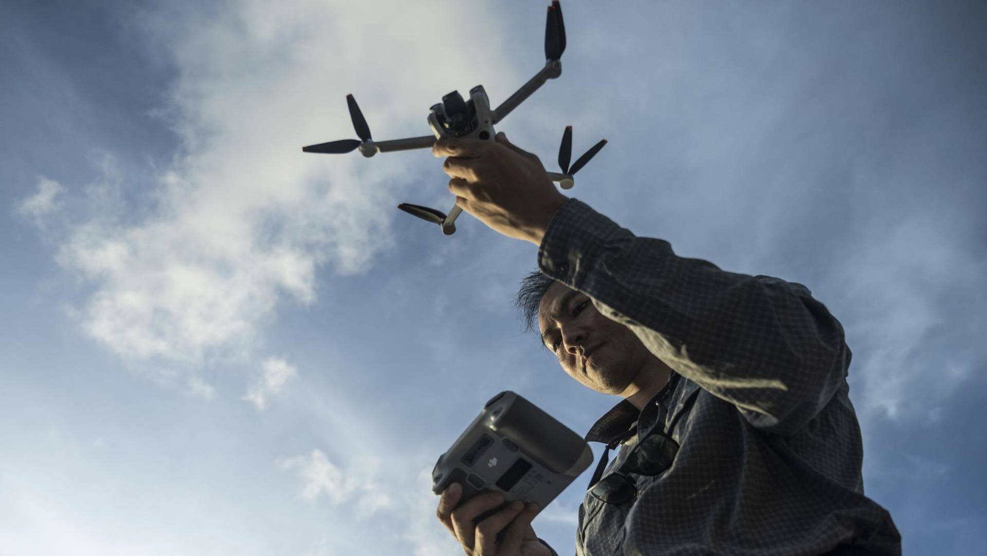 A man holding a drone and drone controller under a blue sky white white clouds. 