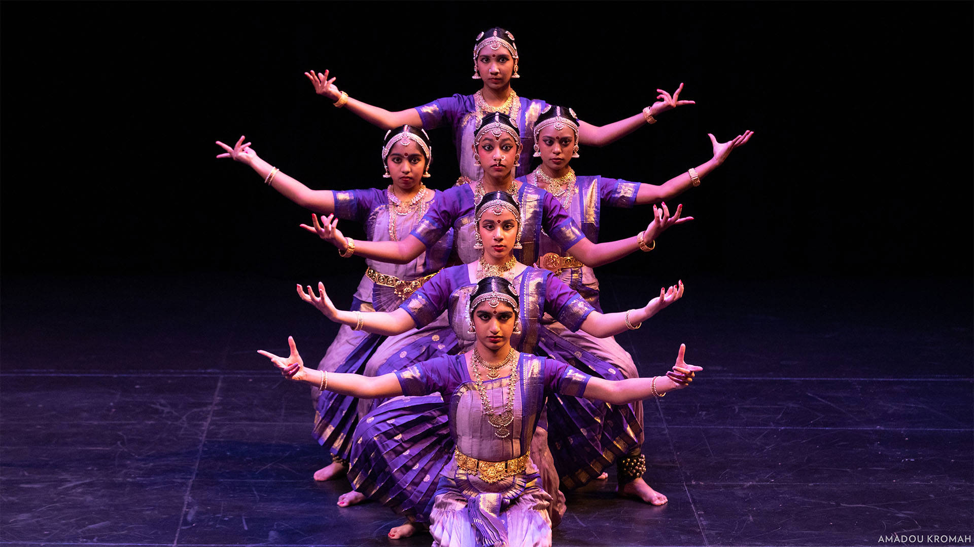 Six dancers in traditional Indian attire perform a synchronized pose on stage.