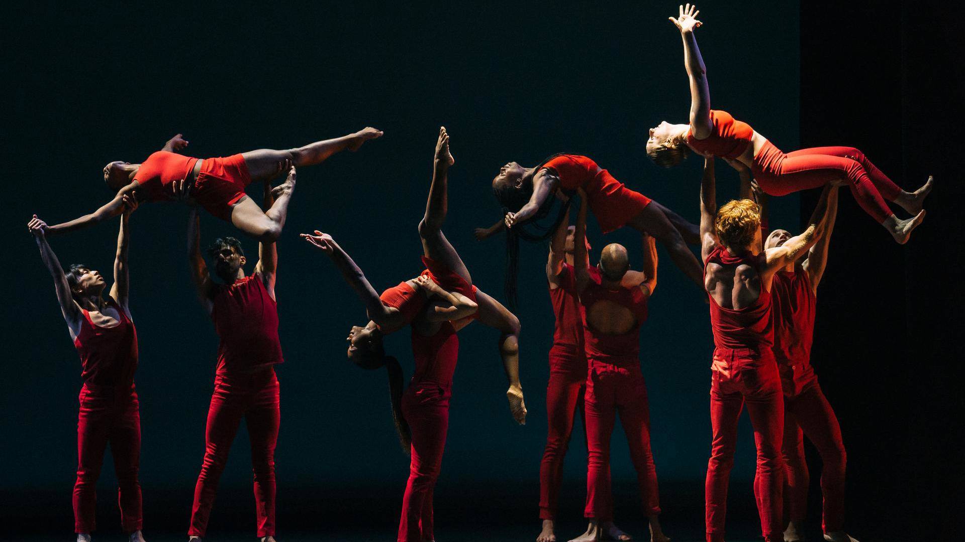 Dancers with light and dark complexions wearing tight red pants and tank tops on a darkly lit stage. Four dancers are being lifted up by pairs of others in graceful poses with arms and legs outstretched.