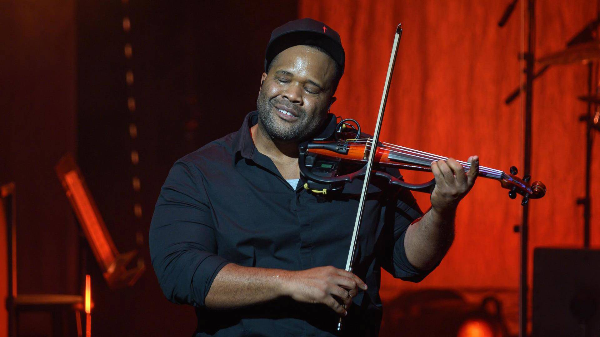 A man with dark complexion, a dark shirt and cap on closes his eyes and smiles as he plays electric violin. The background is red.