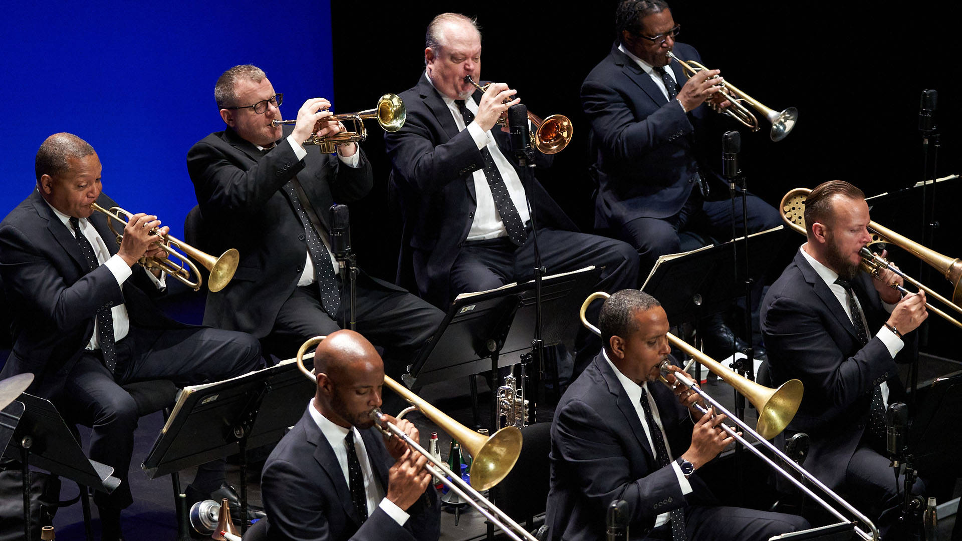 The horn section of the JLCO performing on a bandstand. They are all wearing dark suits.