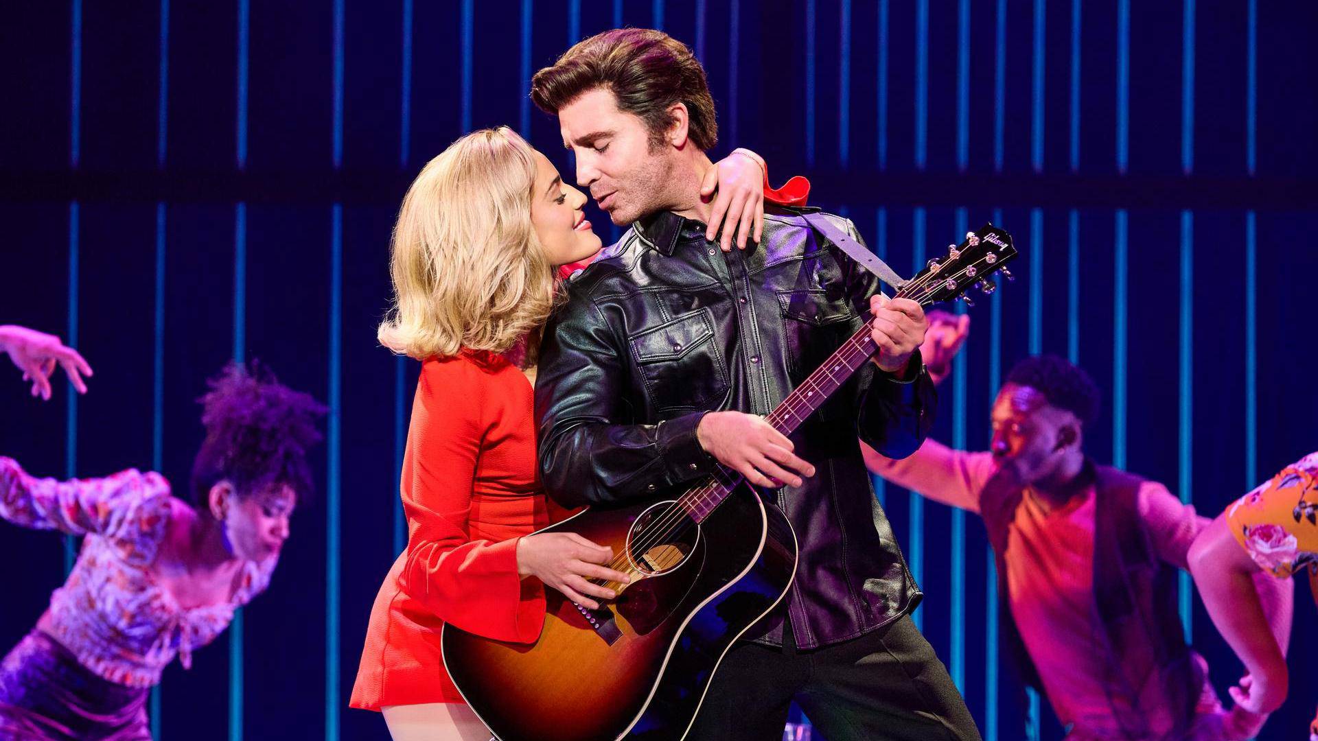 A man with light complexion and greased brown hair in a leather jacket performing with an acoustic guitar. A woman with blonde hair and light complexion in a red dress is draped around him. They are close to kissing.