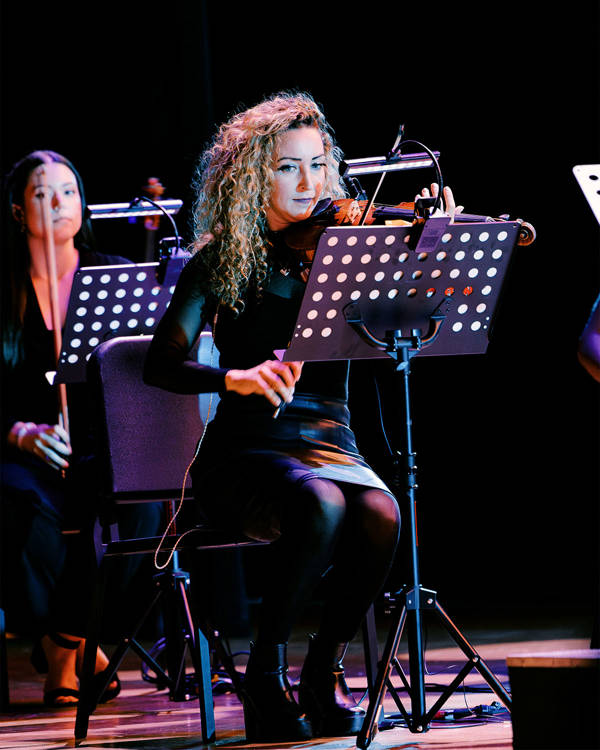 A woman with light complexion, long curly blond hair and a dark outfit playing violin and looking at a music stand.