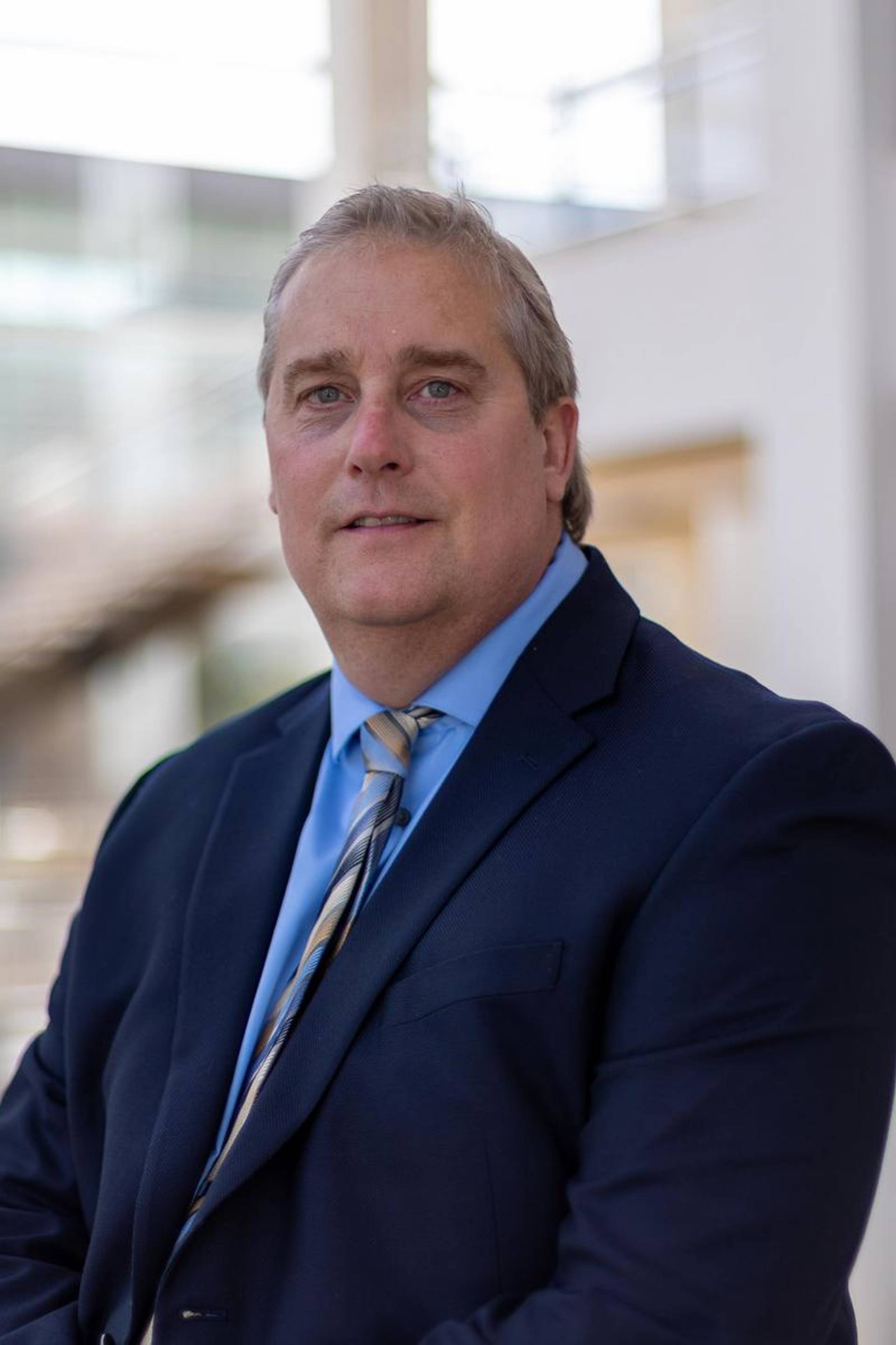 A middle-aged, white man with short, gray hair and hazel eyes. He is wearing a dark blue suit jacket and light blue, button down shirt with a gold and blue striped neck tie.