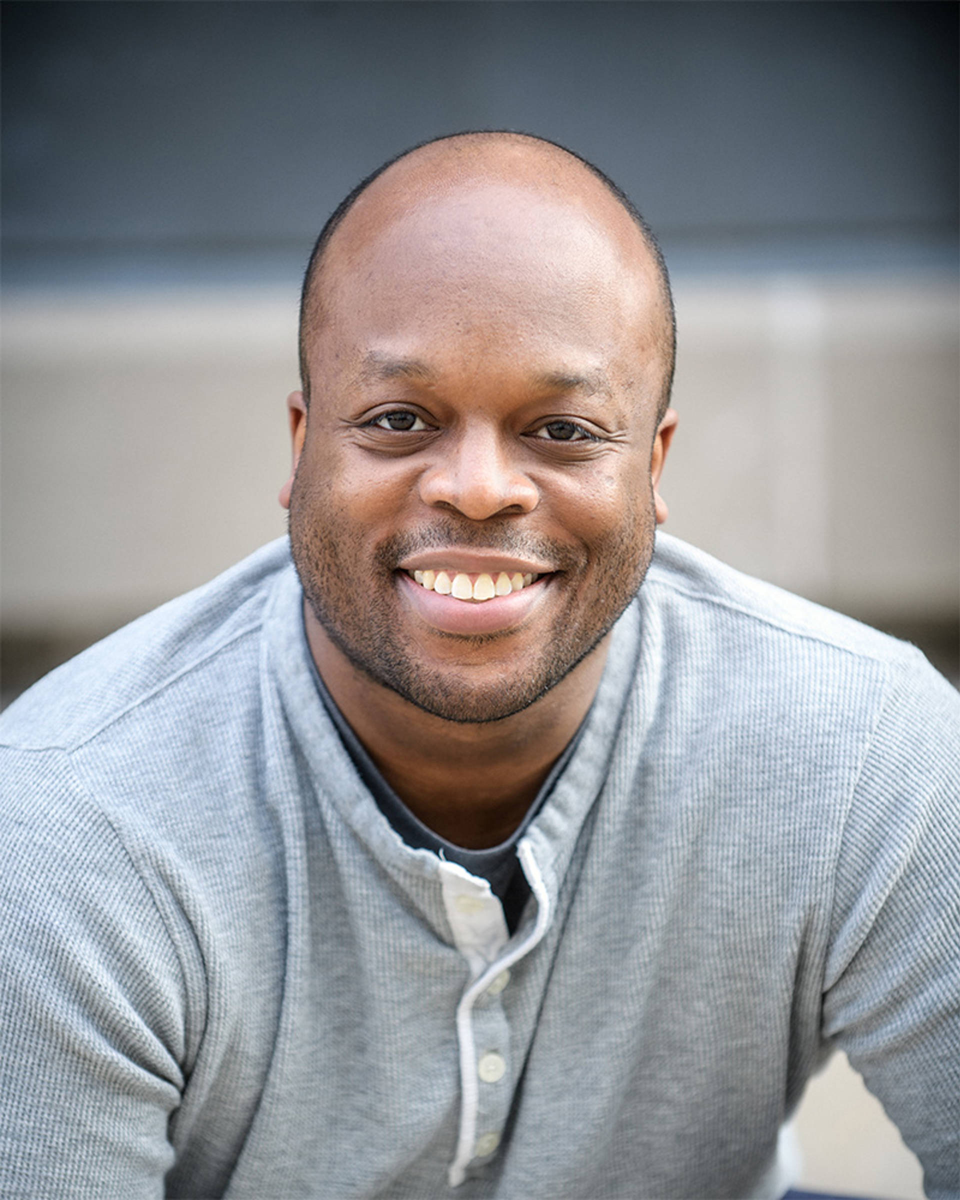 Headshot of a man with dark complexion, dark eyes, a bald head and a grey top.