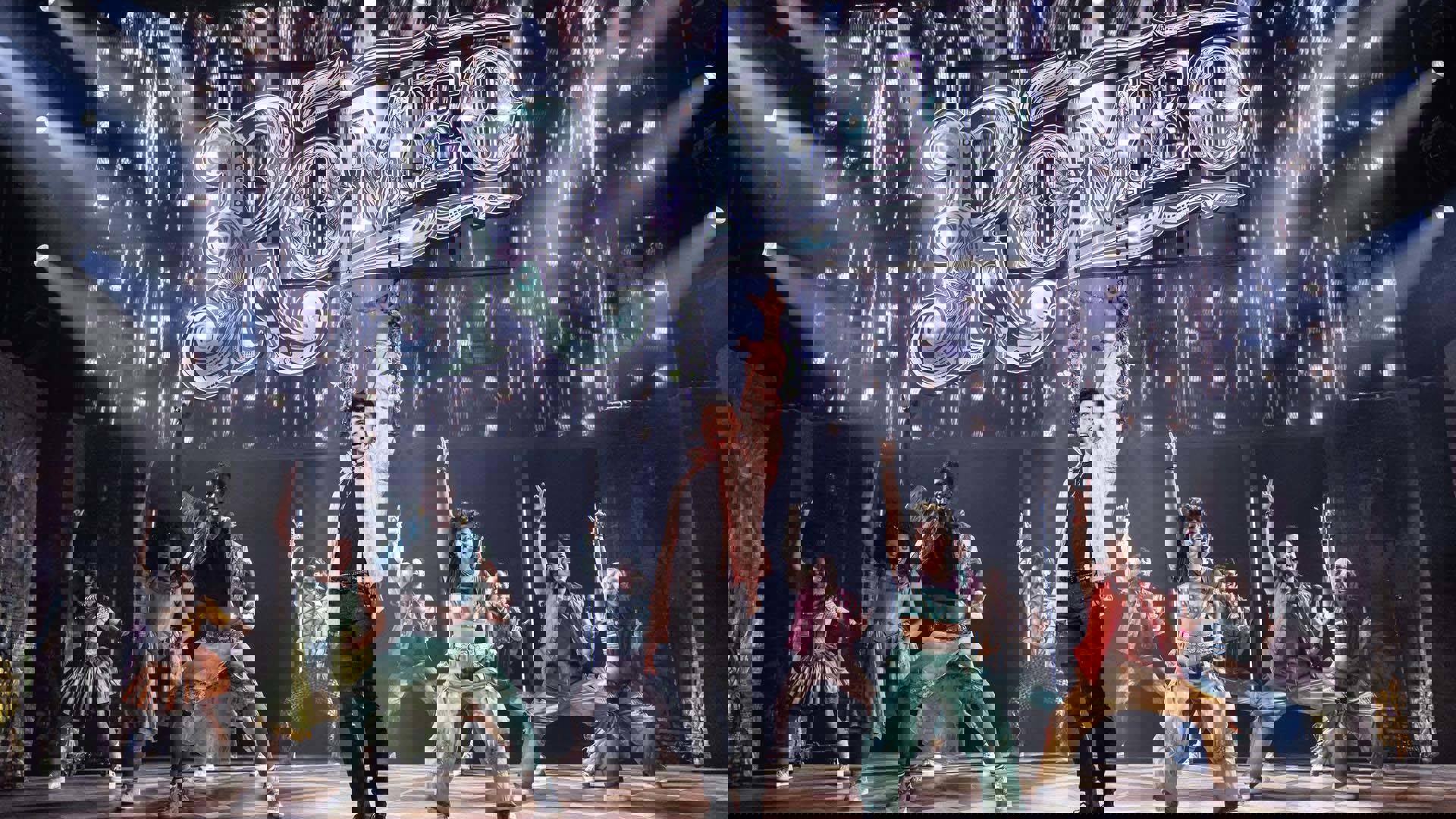 An ensemble performing with their hands in the air in a "rock on" salute. A large silver sign spelling, "Romeo" hangs behind them.