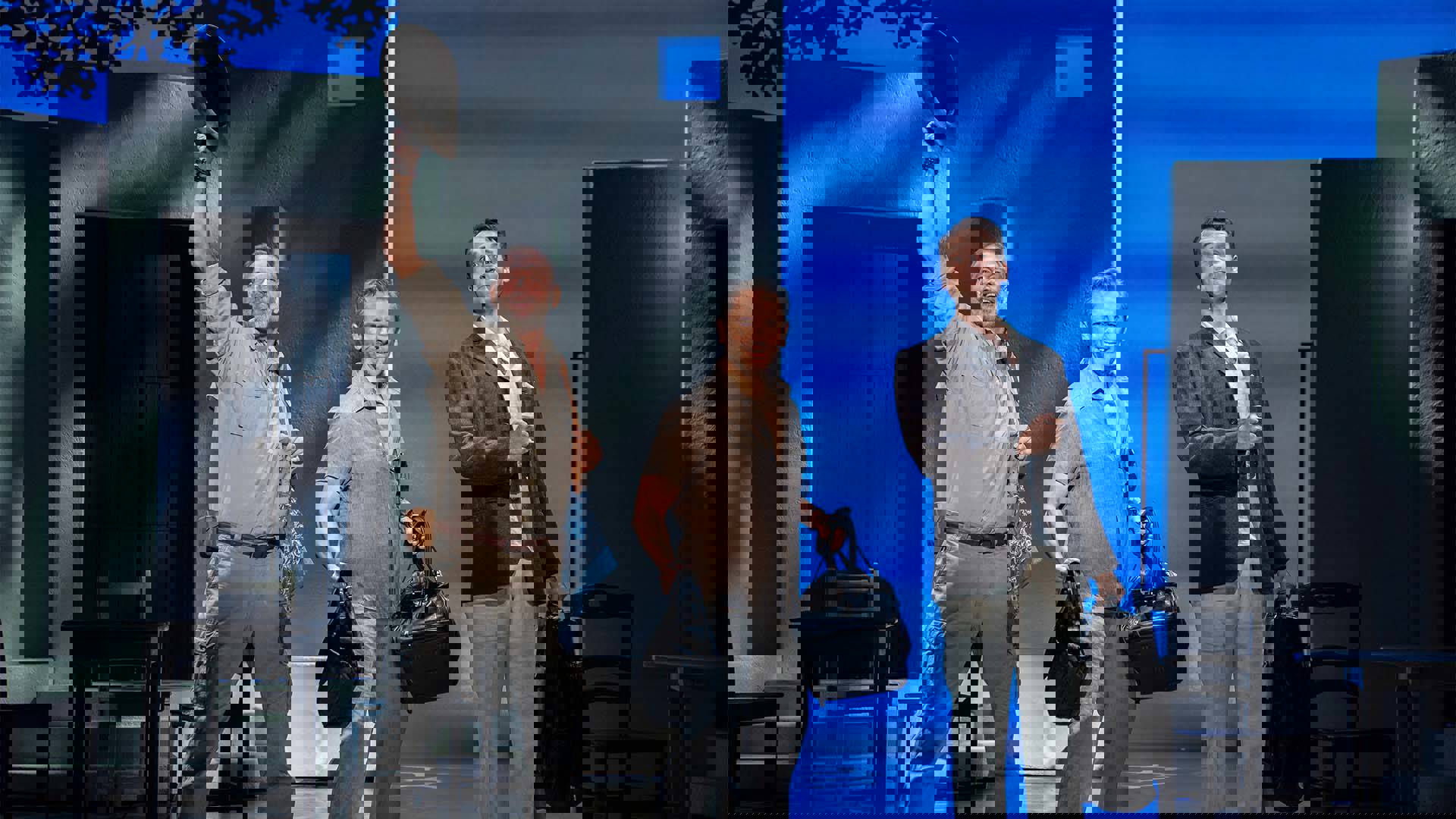 Three middle aged men stand together looking towards the view. They are all carrying bags. The left-most is holding his hat in the air.