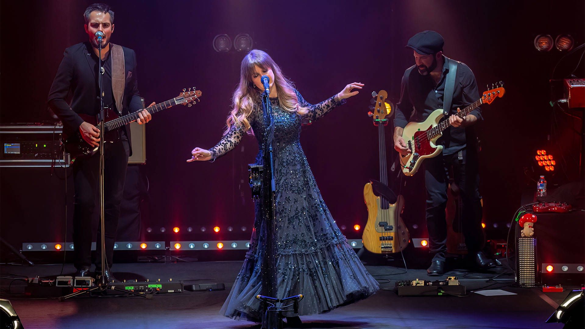 Rumours of Fleetwood Mac band performing on stage. A caucasian woman with long brown-blonde hair stands in the center at a mic stand, singing with multiple spotlights directed at her. She is wearing a longsleeve, glittering, dark blue, flowing long dress and her hands are halfway lifted in the air.