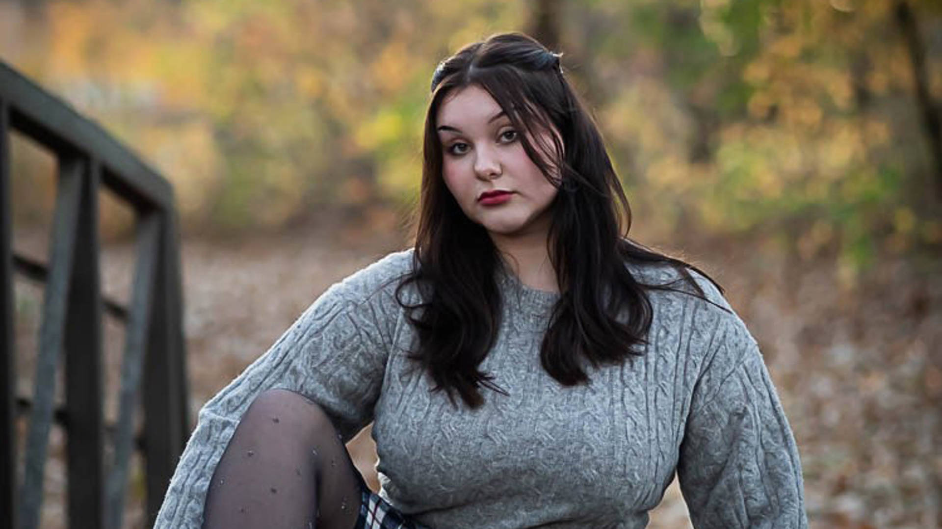 A caucasian high school aged girl sitting for a portrait. She has long dark hair and is wearing a grey sweater and plaid skirt.