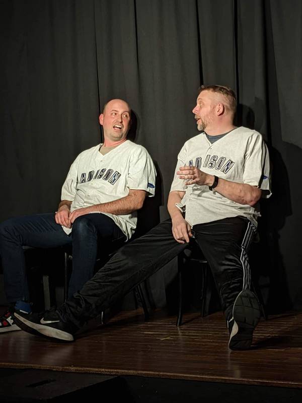 Jess Schuknecht performing improv. He is a white man with short hair and a beard. He is sitgin with another performer, they are both wearing baseball jerseys with "Madison" written on them.