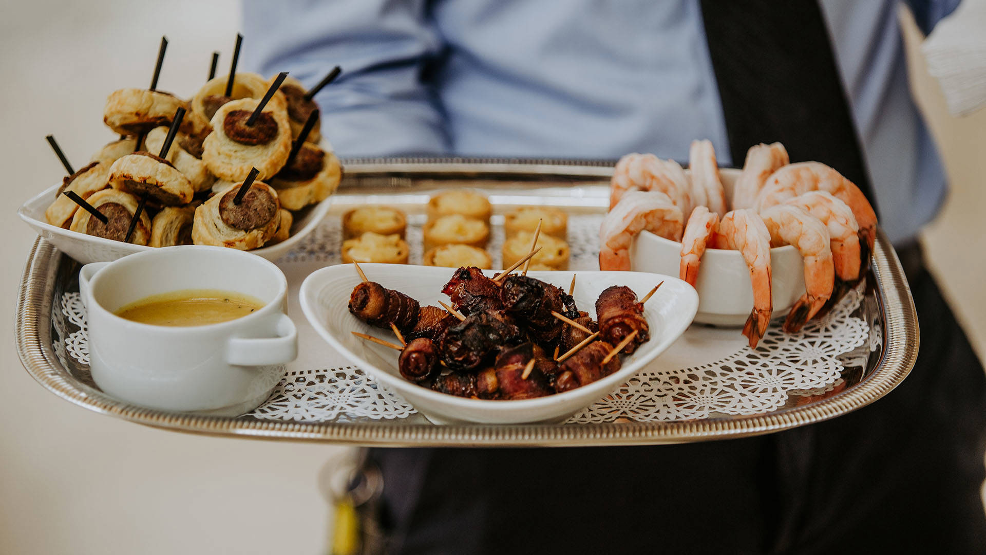 A silver platter with an assortment of hors d'oeuvres, including shrimp cocktail, bacon wrapped dates, pigs in a blanket, and little bites, held by a person in a blue dress shirt and black tie.