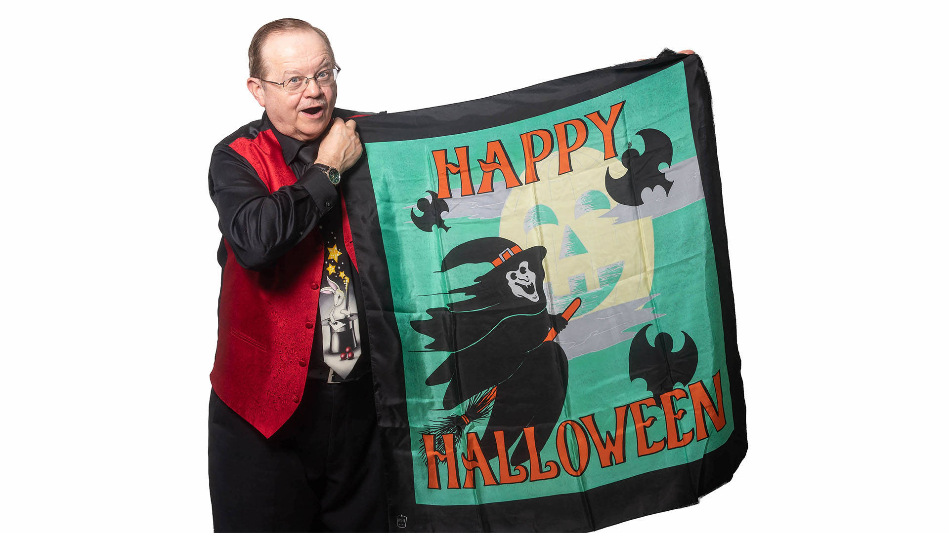 An older caucasian man with receding brown hair and glasses holds up a "Happy Halloween" sign. He is wearing a black suit with a red vest and tie the depicts a rabbit in a magic hat.