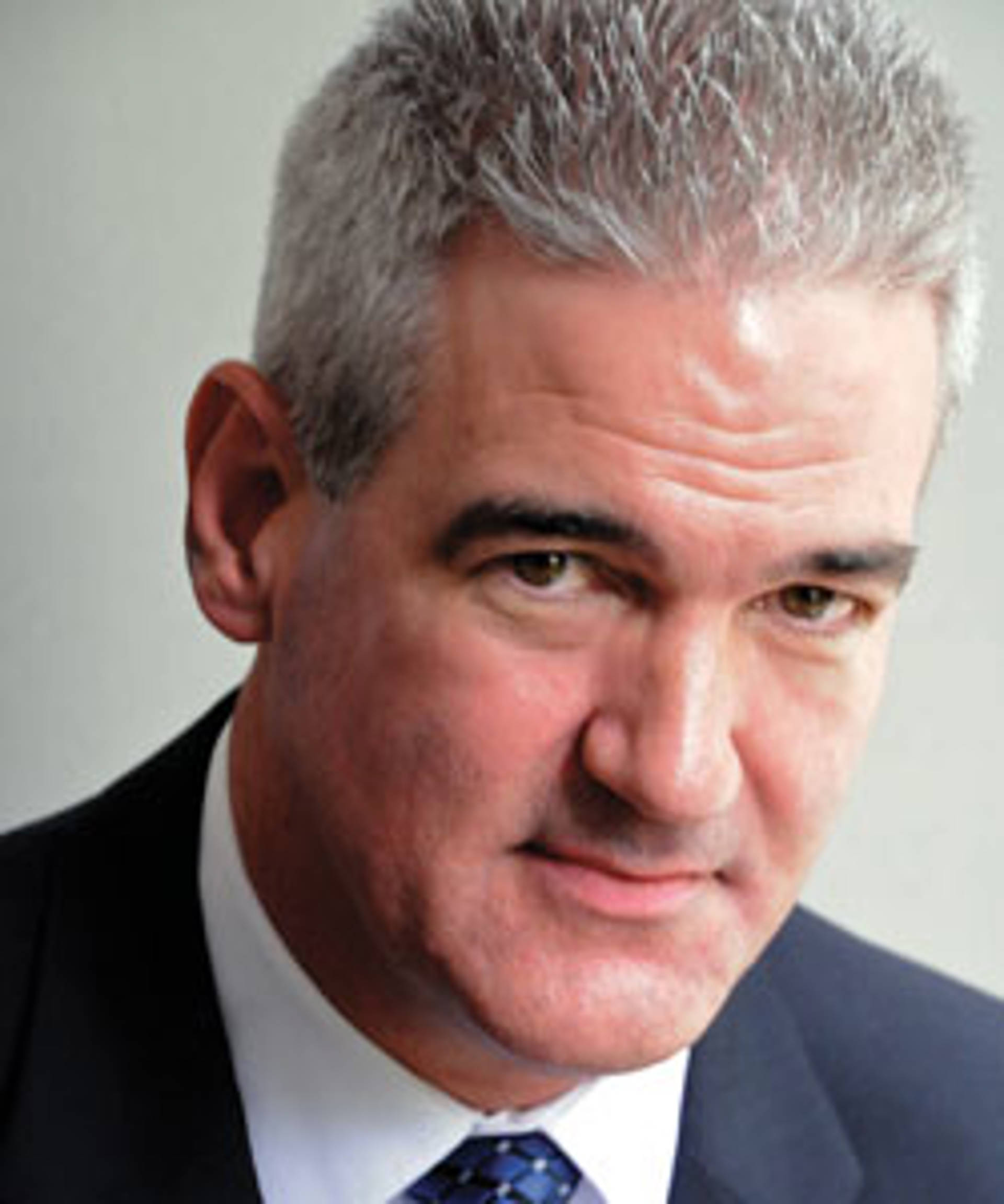 Headshot of a caucasian man with brown eyes short grey hair. He is wearing a dark blue suit jacket and tie.