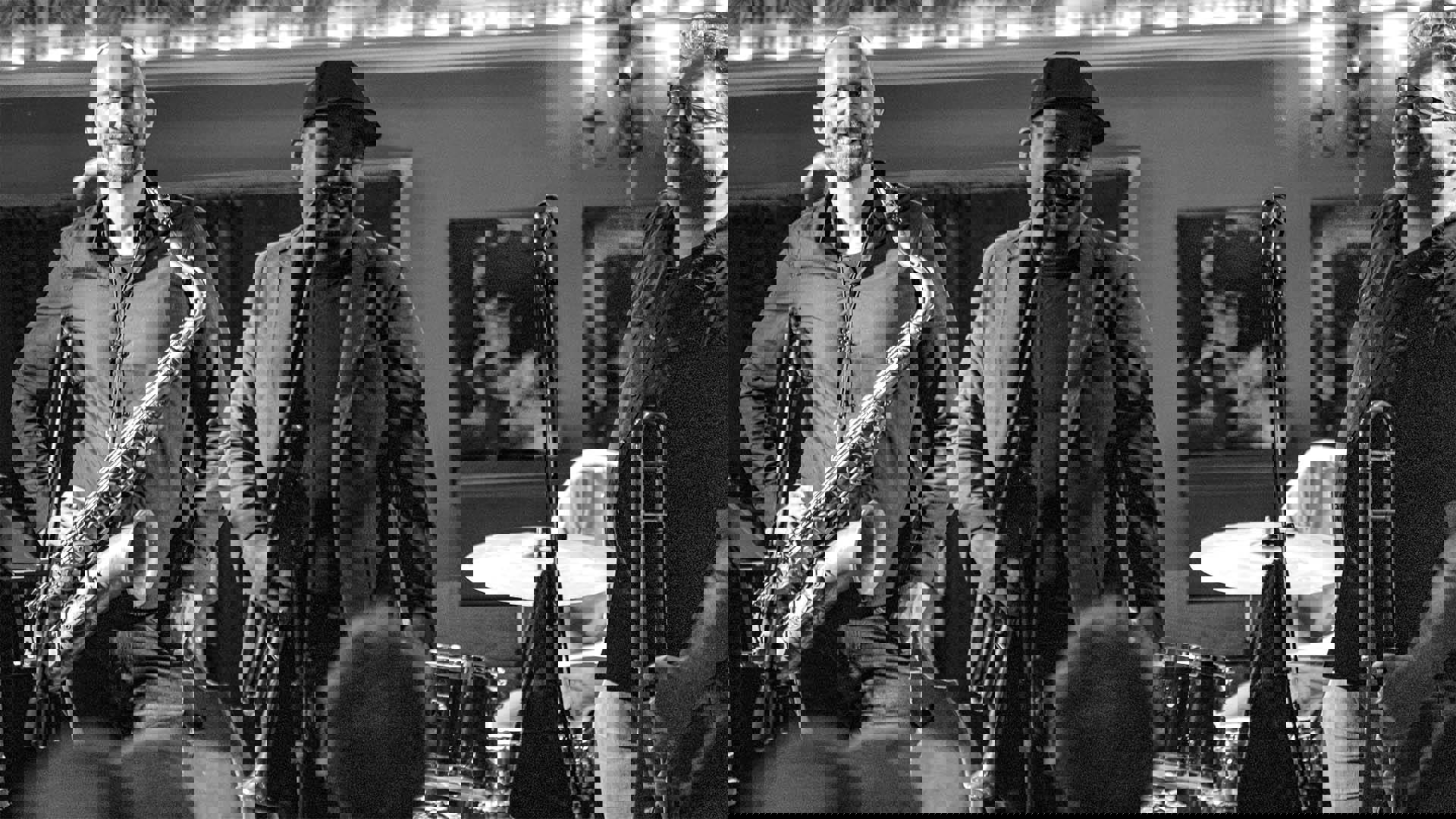 Three men performing jazz with a saxophone, trumpet and trombone and a drum kit behind them. Black and white image.