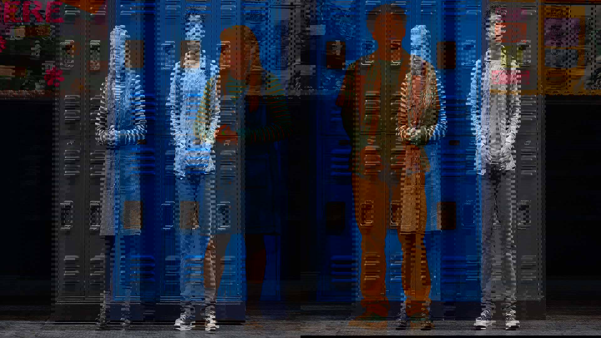 A woman with long hair and light complexion, and a man with medium to dark complexion and short, dark hair stand looking concerned in front of a wall of blue lockers.