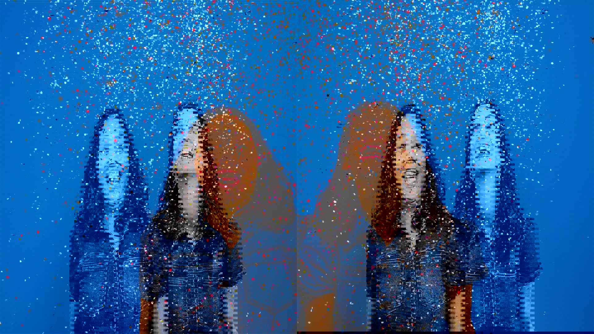 Two women laughing in matching jean tops against a blue background with the confetti falling all around them.