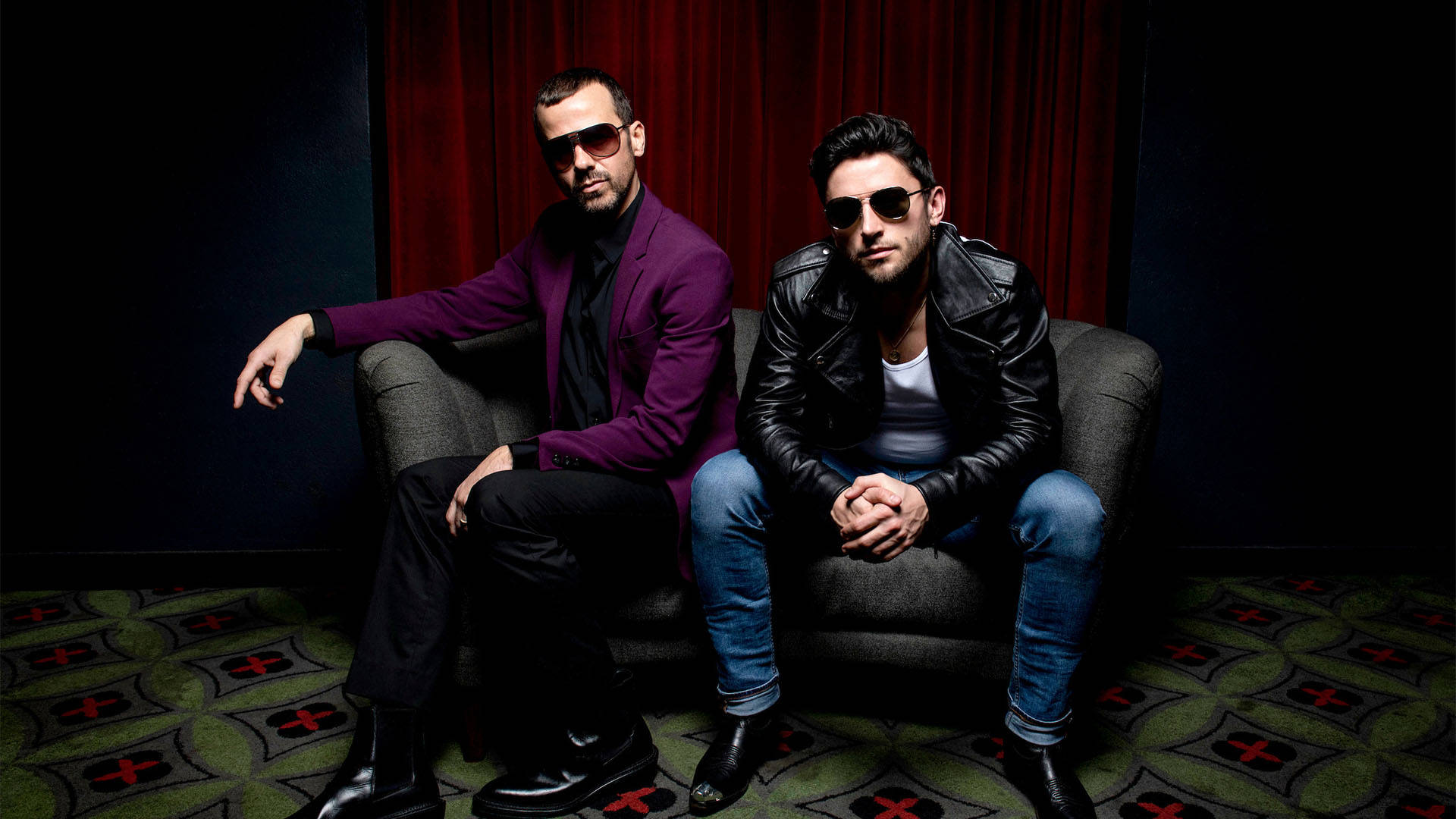 Two men sitting together on a small couch posing for a photo. The man on the left has short brown hair, sunglasses and a purple jacket. The man on the right has short brown hair, jeans and a leather jacket.