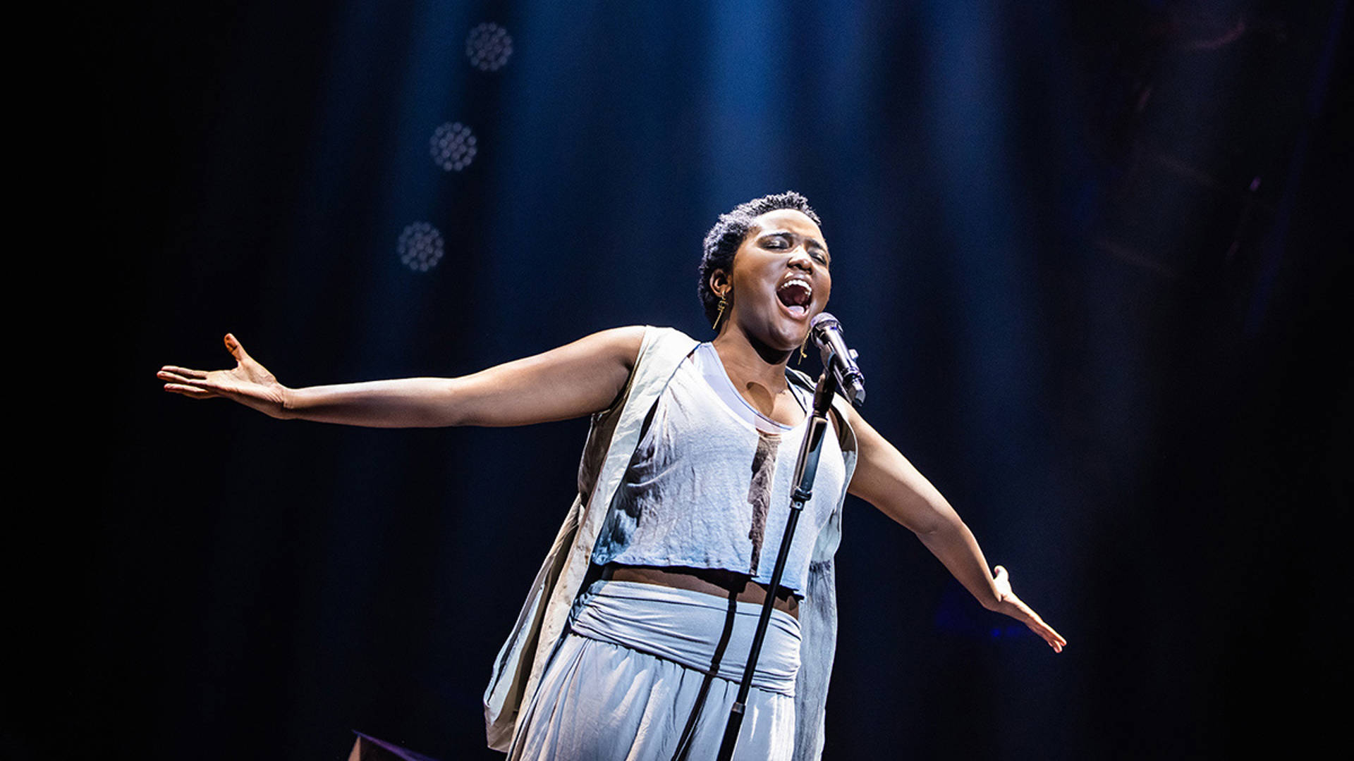 An African American woman with short hair and white clothing sings with arms stretched wide.