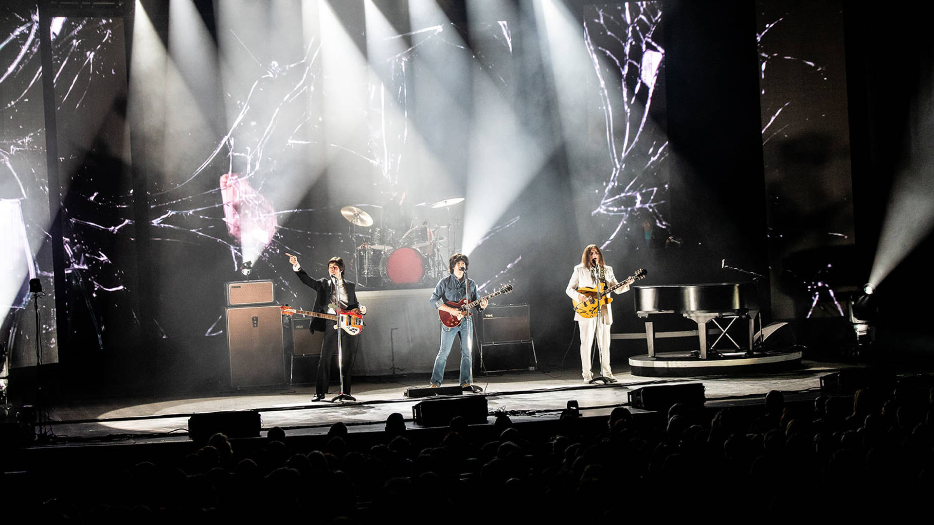 Four Beatles impersonators performing on a brightly lit stage, wearing stylish 1960s street clothes and holding guitars.