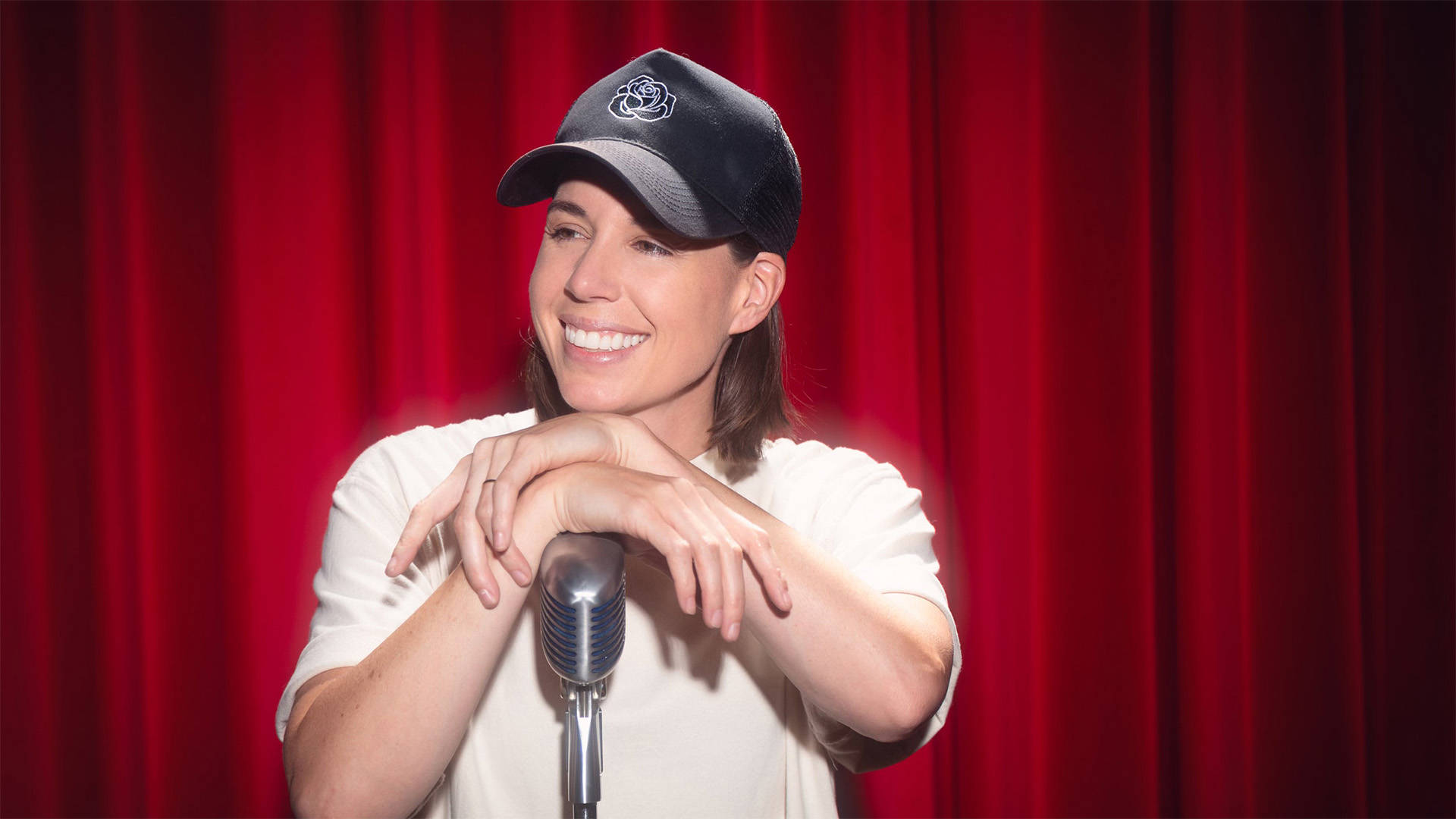 Danae Hays, a woman with light complexion, medium length brown hair, a baseball cap and a white tshirt, smiling and resting her arms on a microphone on a stand.