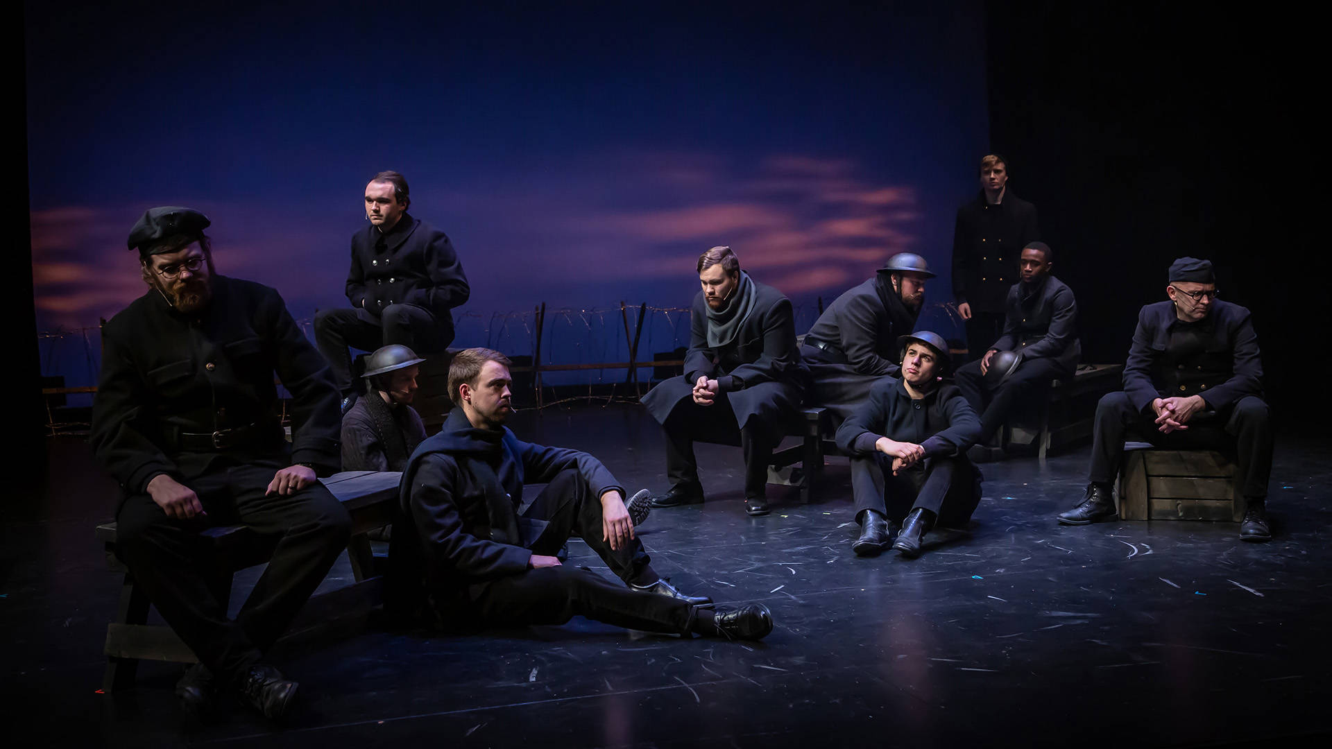 Ten actors in WW1 soldiers outfits sitting with pensive looks on their faces in a dark space.