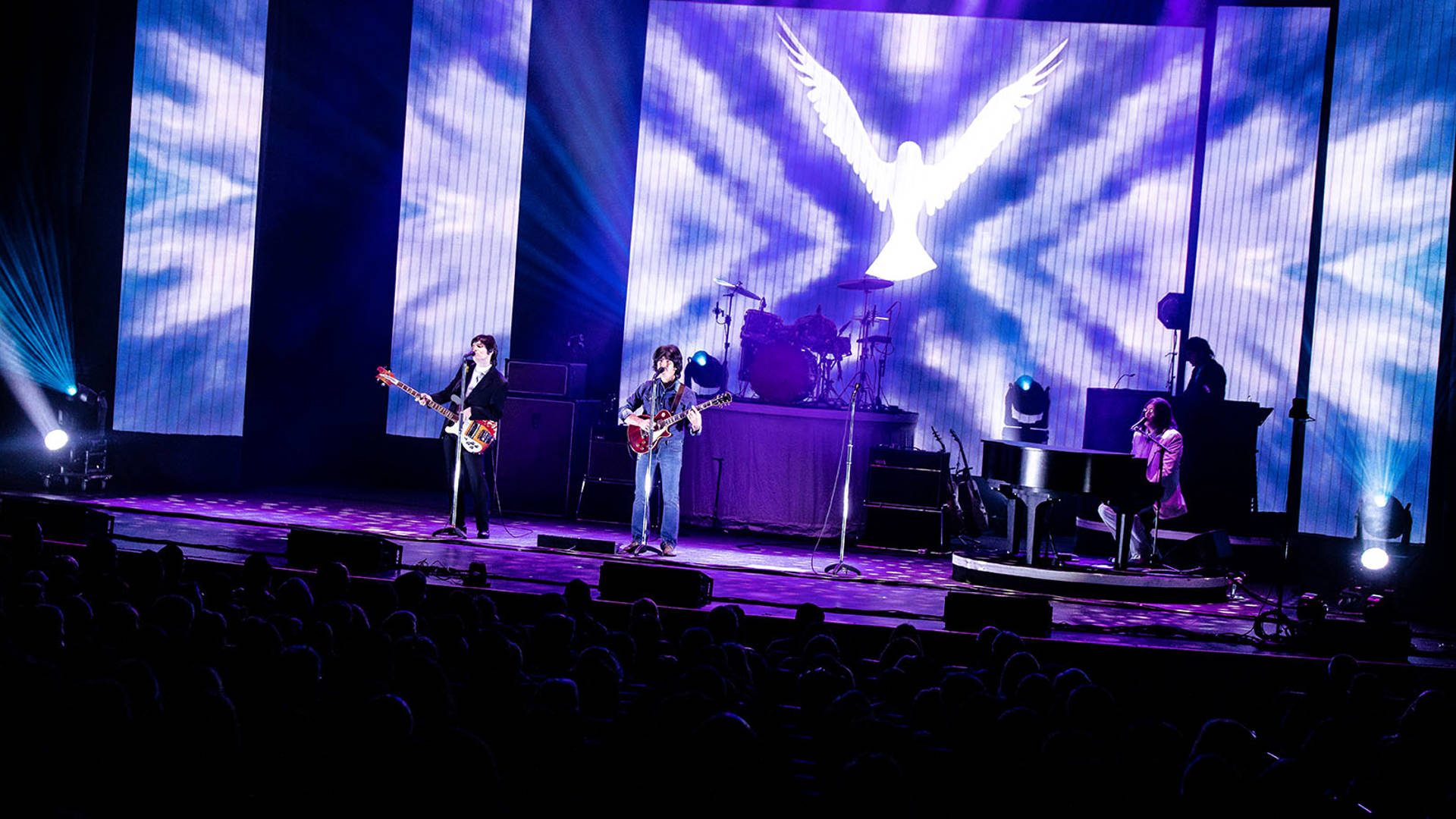 Four Beatles impersonators performing on a dark blue stage, wearing stylish 1960s street clothes and holding guitars.