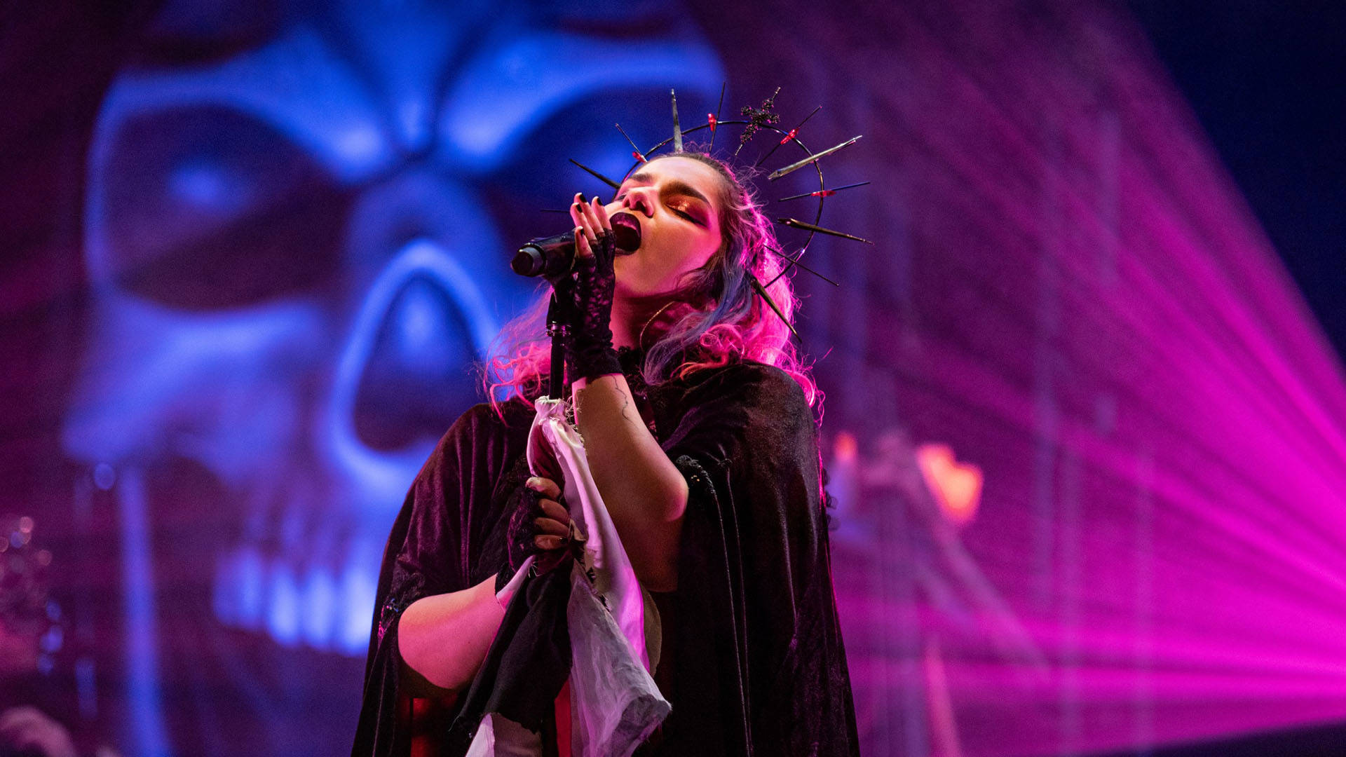 Woman wearing gothic crown and black cape sings into a microphone with large skull and pink light beams in background.