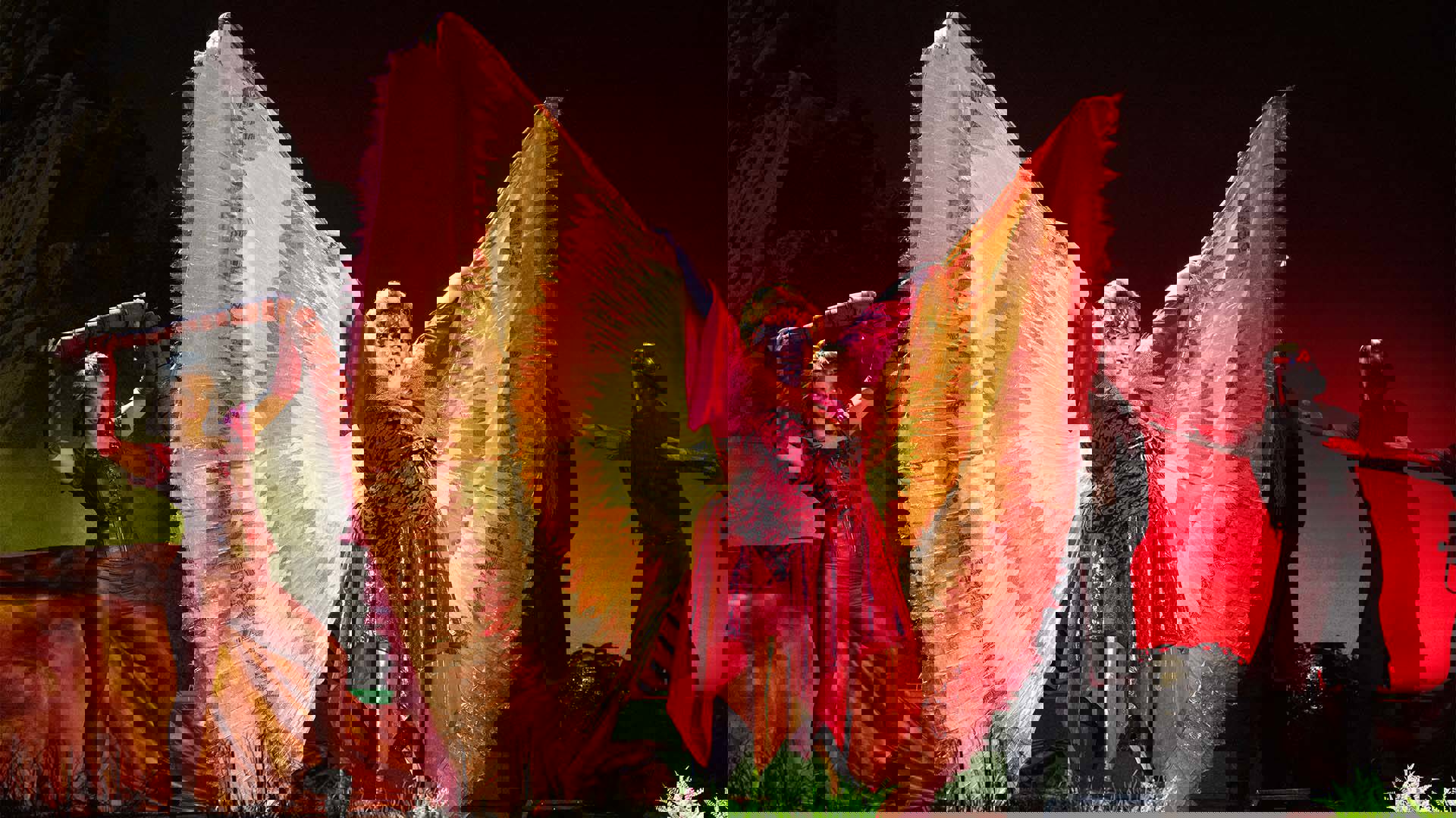 A caucasian woman dressed as Dragon. She is wearing a red shiny layered dress with her arms raised up, displaying wings of yellow and orange fabric.