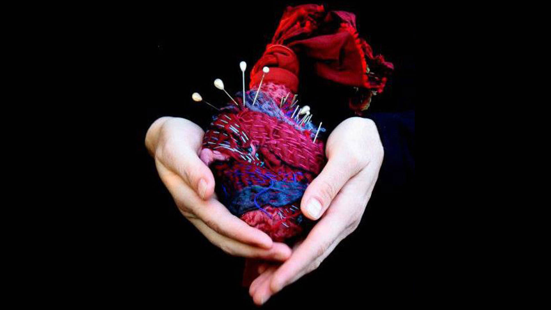 A pair of caucasian hands holding a red, purple and blue yarn heart with white pearl pins sticking into the top. Sculpture by Lorraine Reynolds.