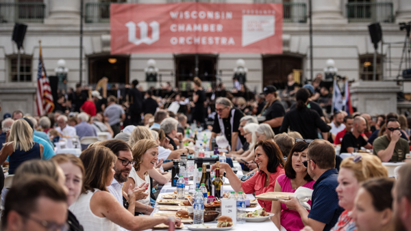 Concert goers enjoy dinner at a long table in front of the Wisconsin capitol building.