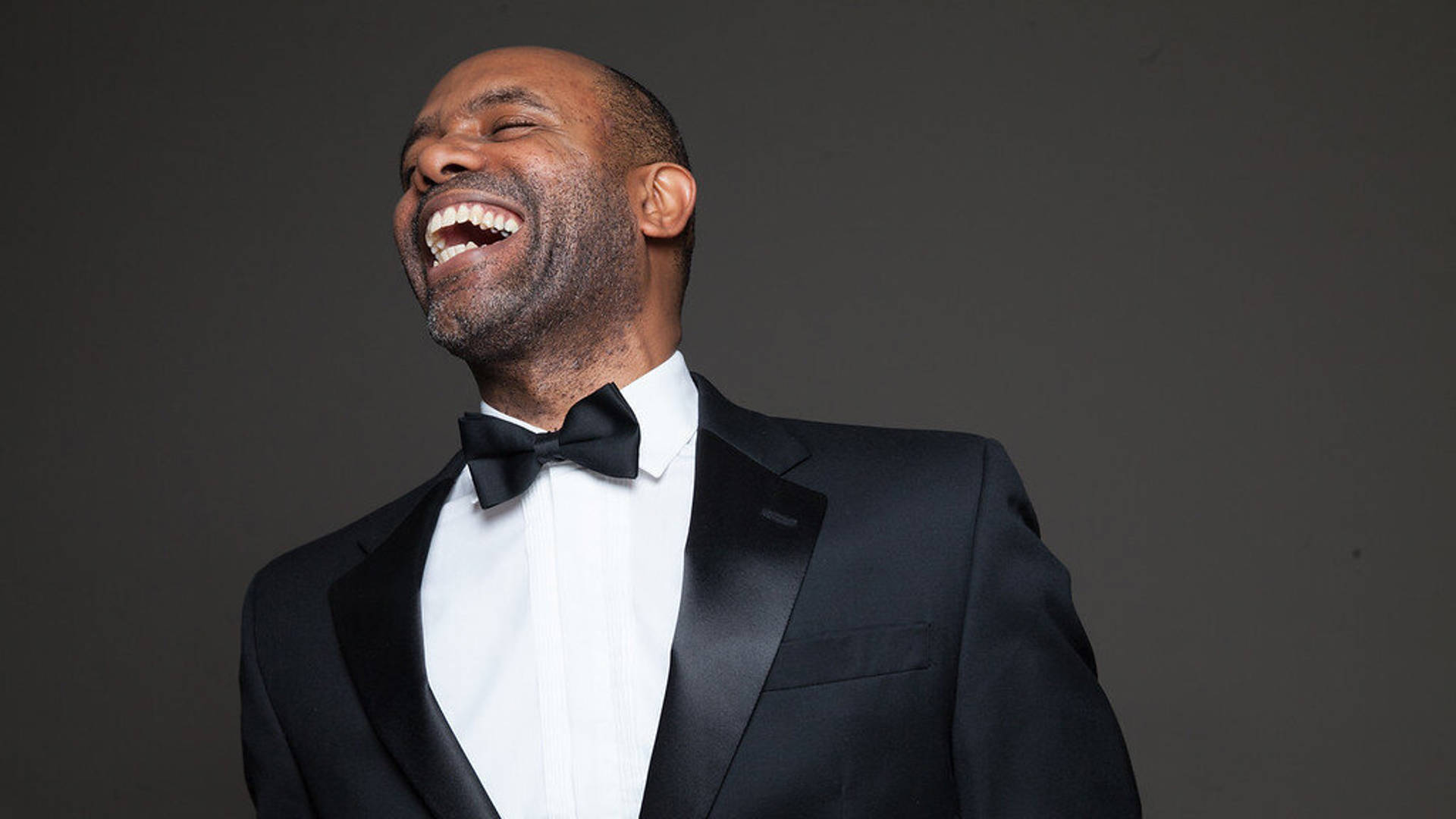 An African American man with short dark hair wearing a tuxedo. His eyes are closed as he laughs heartily.