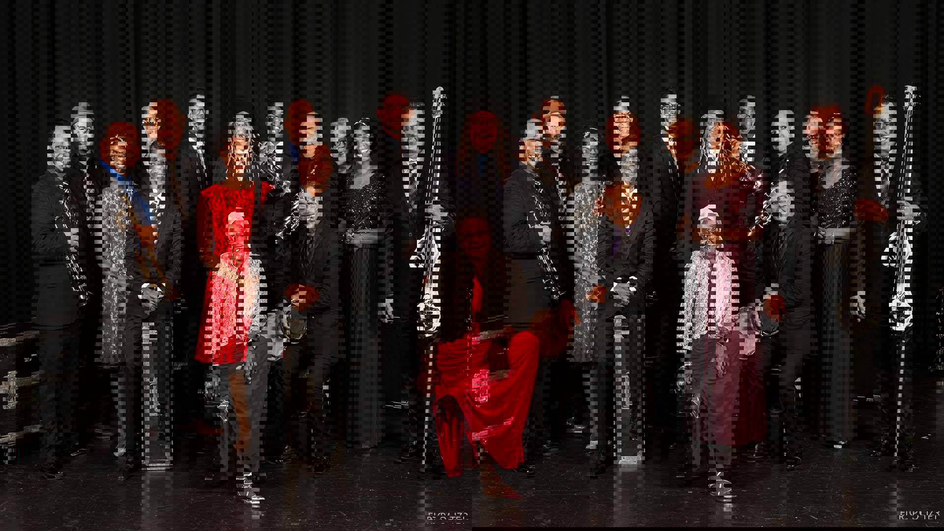 The 14 members of the Afro-Cuban Allstars stand together holding a variety of instruments. They are all dressed beautiful suits and dresses.