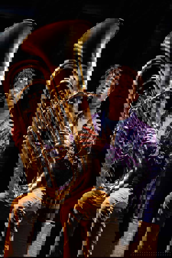 A man with medium to dark complexion and dark, curly hair sits and plays a tuba.