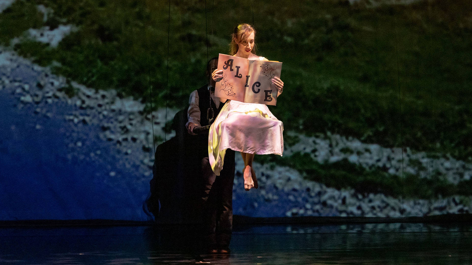 A dancer with long brown hair in a ponytail wearing a white shiny dress is floating on stage reading a large book that says "Alice" across the cover.