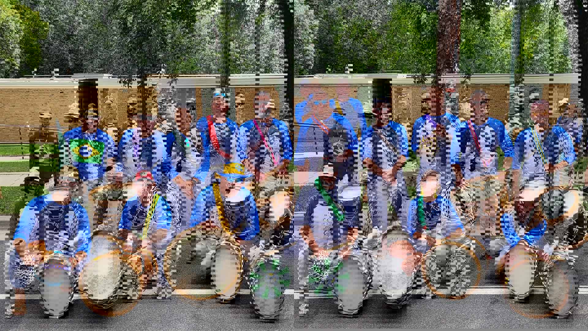 A smiling group of drummers outside showing off a variety of brazilian drums. They are all wearing matching blue and white baseball Tshirts.