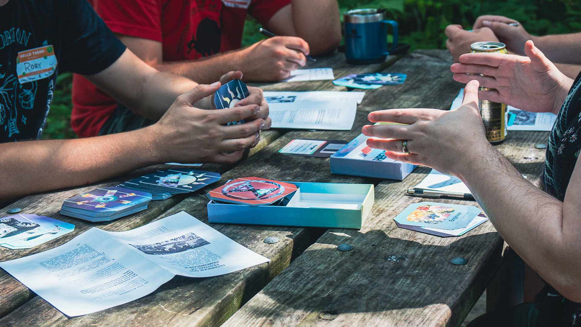 Four people playing Solarpunk Futures on a wooden picnic table outside. The image only shows people's hands with game cards laid out on the table.
